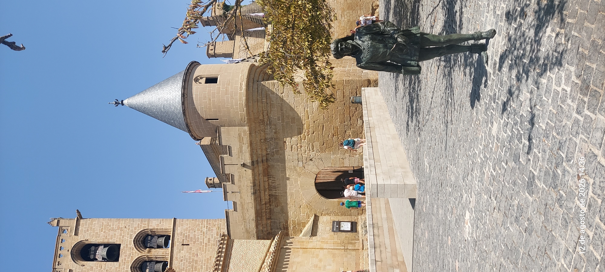 The image shows a courtyard in front of a stone building with a conical roofed tower. A bronze statue of a man in historical attire stands in the foreground on a cobblestone ground. In the background, people are walking through a large arched doorway into the building. A bell tower with three bells is visible on the left side of the building. The sky is clear and blue. The timestamp at the bottom of the image indicates "12 de agosto de 2020 14:36". This location is the Palace of the Kings of Navarre in Olite, Spain.