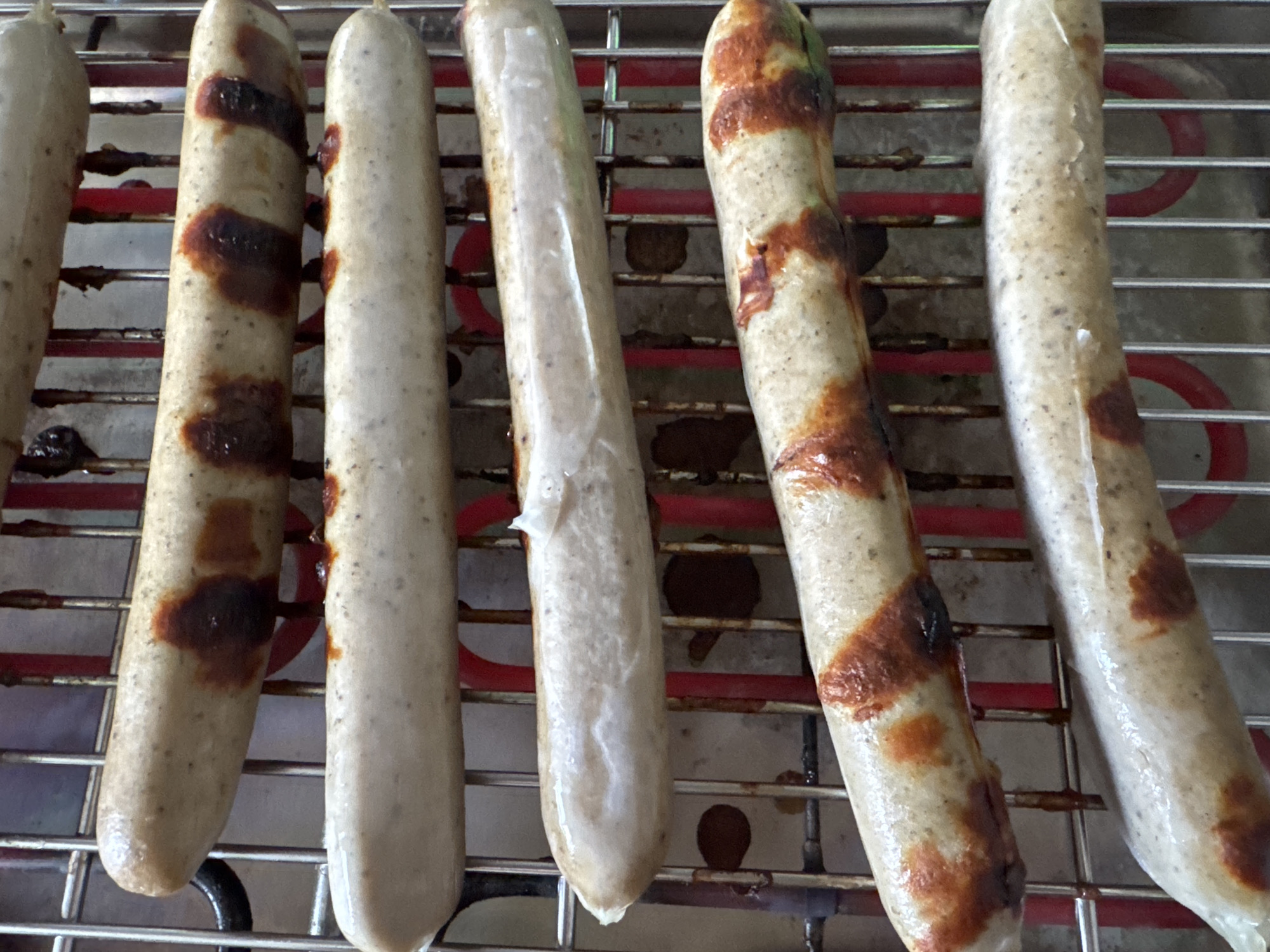 The image shows five sausages cooking on a grill. The sausages are arranged horizontally in rows on a metal grate. The grill appears to be electric, with red heating elements visible underneath the grate. Some of the sausages have grill marks, indicating they are actively cooking. The background is blurred, but suggests an indoor setting. The image was taken in Höhenkirchen-Siegertsbrunn, Germany.