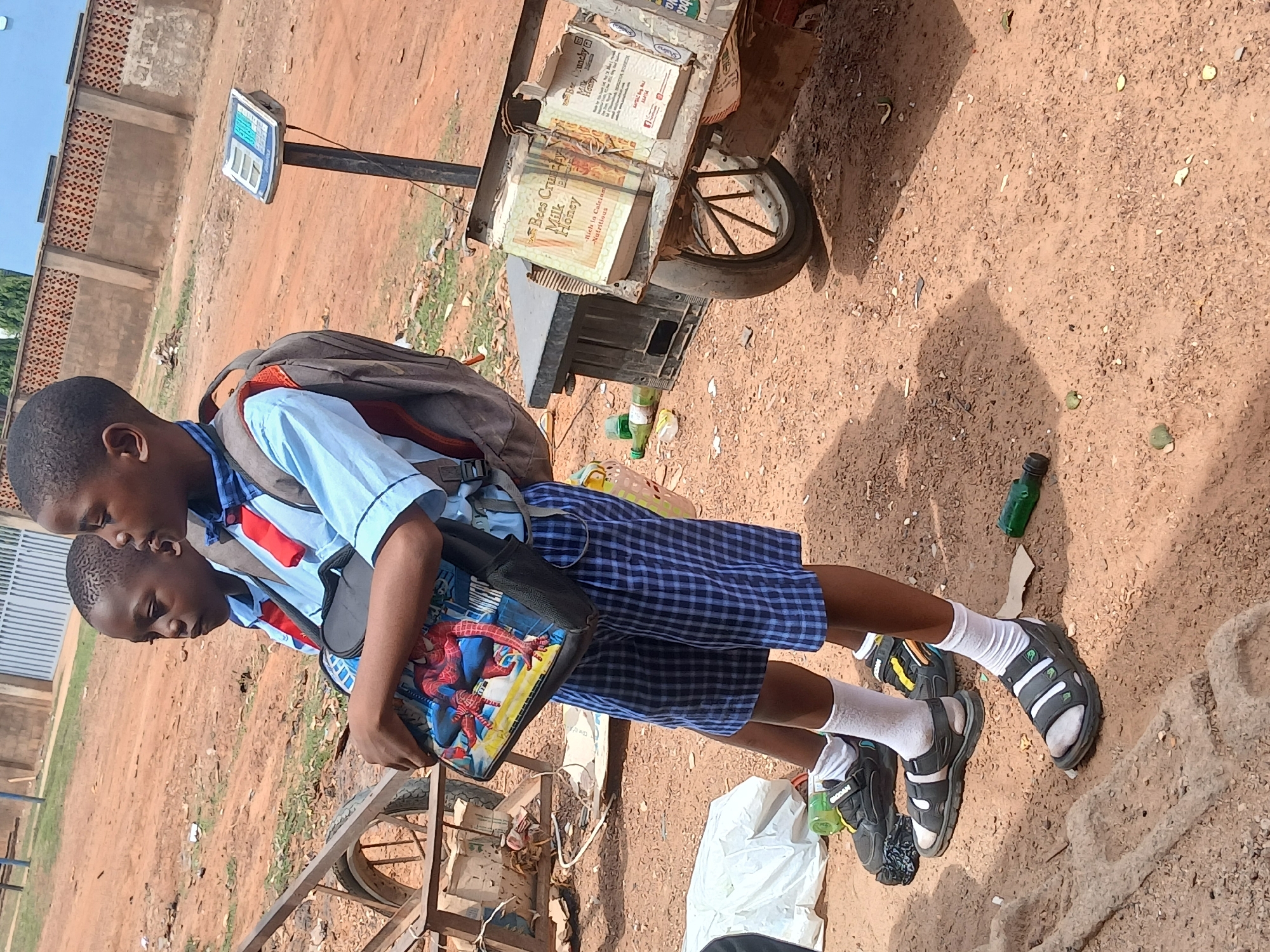 Two young boys in school uniforms stand next to a cart in Rido, Nigeria. The boys wear light blue shirts, red ties, and blue plaid shorts. The older boy has a grey backpack and a bag with a Spiderman design. They both wear white socks and sandals. The cart contains books with the label "Bees Crush Milk Honey" visible. A digital scale stands near the cart. The ground is reddish-brown soil with scattered debris, including plastic bottles. A low brick wall is visible in the background.