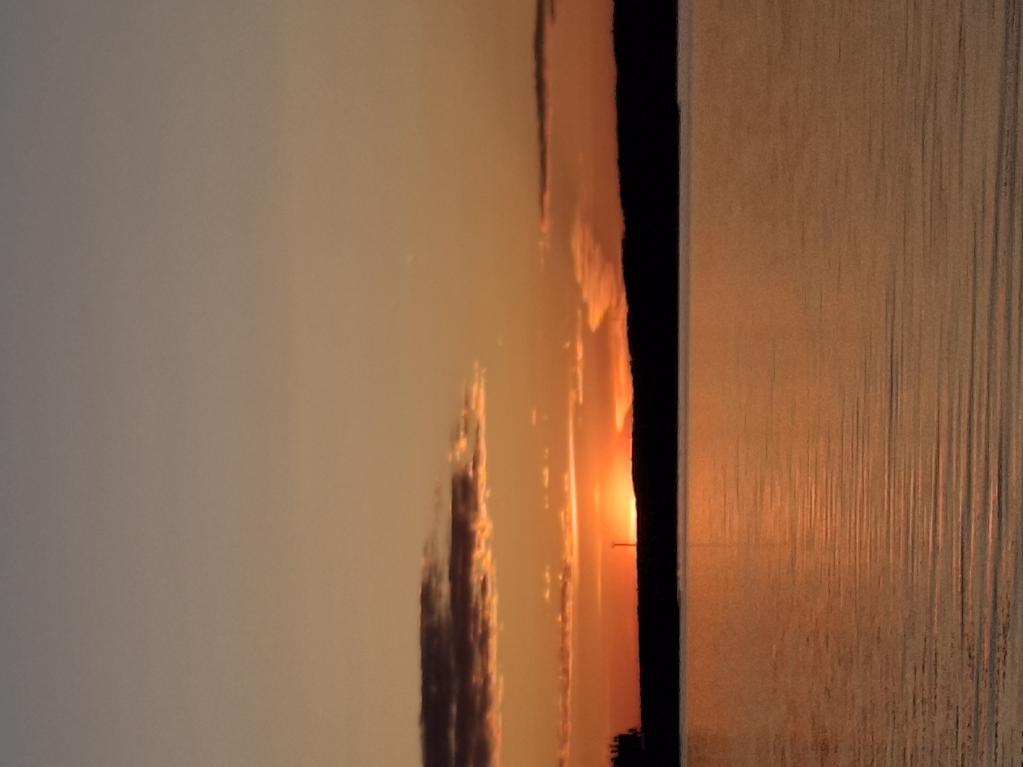 The image captures a sunset over the Kama River in Chaikovsky, Russia. The setting sun casts a bright orange glow on the water's surface, creating shimmering reflections. Silhouettes of trees and buildings form a dark line along the horizon. The sky displays shades of orange and gray, with scattered clouds.