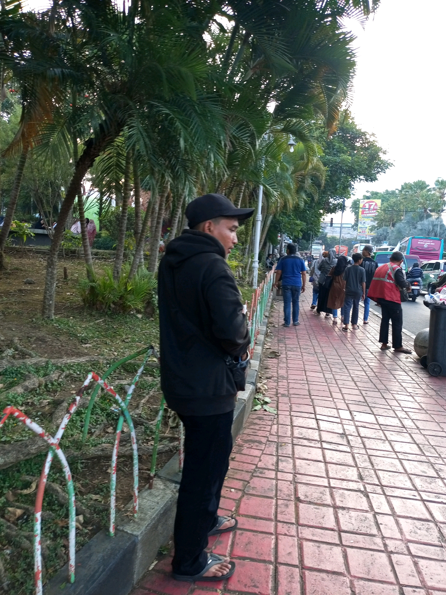A medium shot captures a man in a black hoodie and black pants standing on a red brick sidewalk, facing away from the camera and towards a street scene. He is wearing black flip-flops. The man's left hand is visible, and he appears to be looking to his right. The sidewalk is lined with green grass and palm trees on the left. In the background, several people are walking on the sidewalk. To the right, a street with traffic is visible, including buses, cars, and a motorcycle. A large black trash bin with wheels is situated on the sidewalk to the far right. The scene appears to be in Batu, Indonesia, possibly near a park or recreational area, given the presence of trees and greenery. The overall lighting suggests it is daytime.