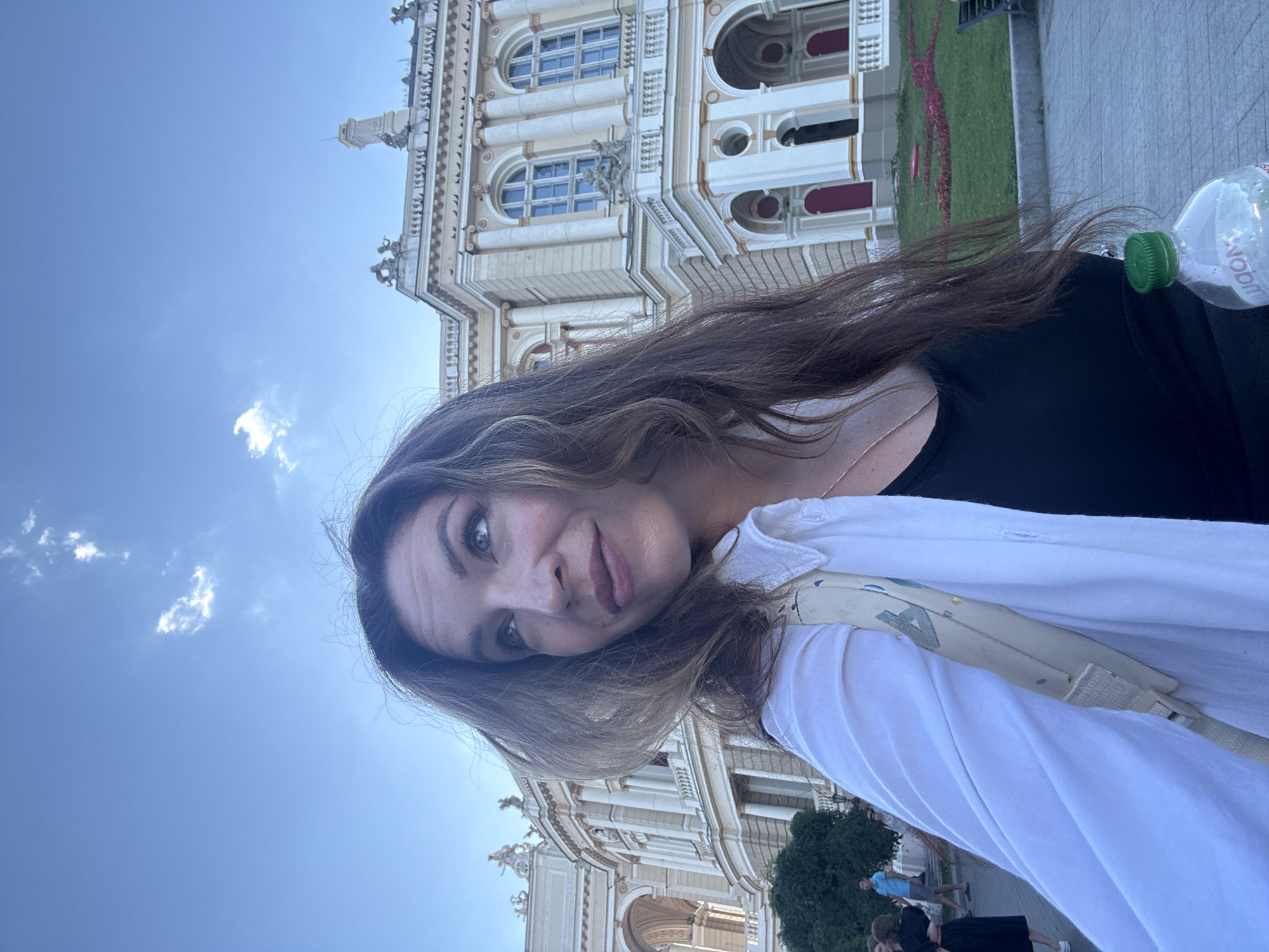 A woman is taking a selfie outdoors with a building in the background. The building is an ornate, light-colored structure with multiple arches, columns, and windows. It appears to be a historic or public building, possibly a theater or opera house. The woman is wearing a white open shirt over a black top, and she has long, brown hair. She is holding a clear plastic bottle of water with a green cap. In the background, other people can be seen walking and standing, and there is a grassy area with some landscaping. The sky is blue with a few white clouds. The scene is likely in a city square or park in Odesa, Ukraine, given the architecture and setting.