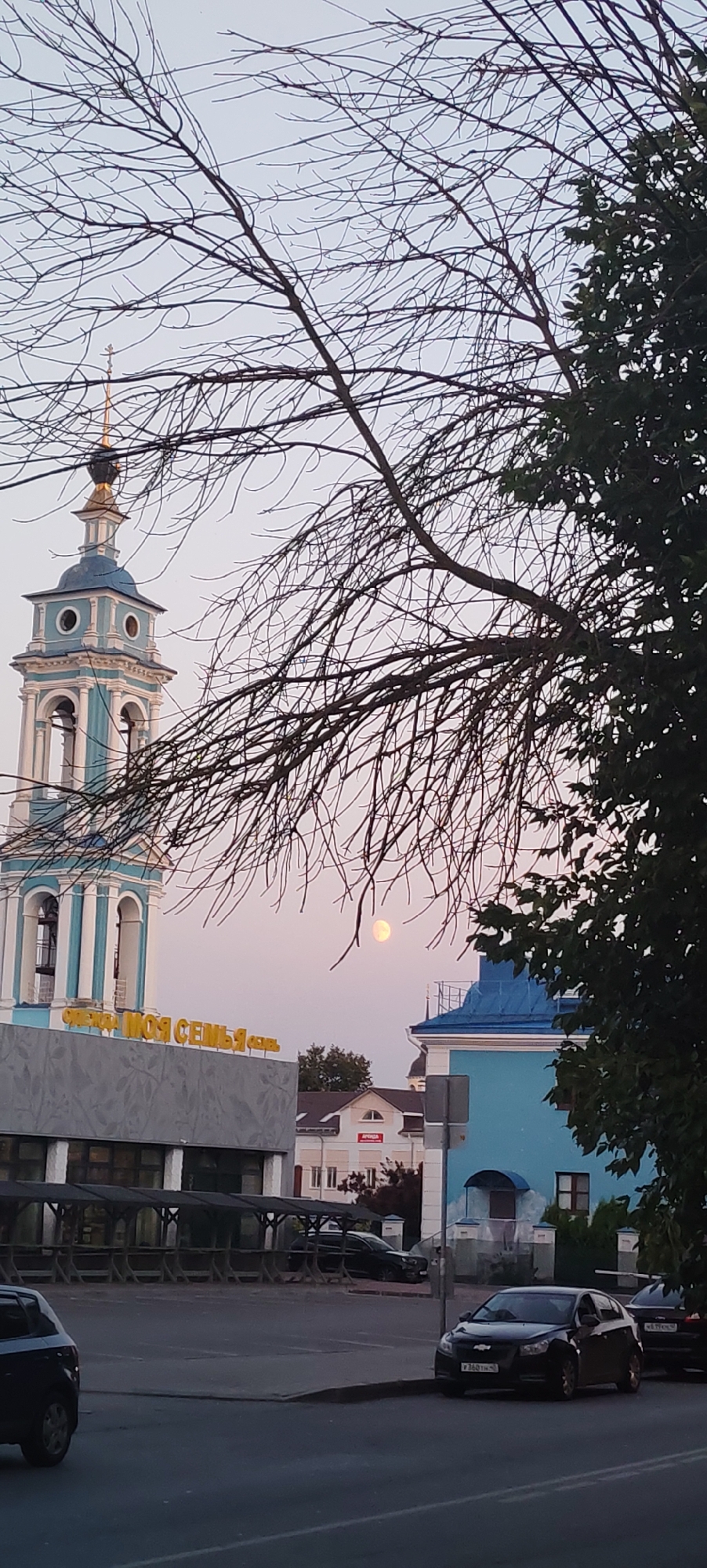 A view of the Church of St. Nicholas the Wonderworker in the city of Kaluga, Russia. The church's blue and white bell tower stands tall against a soft, pink sky, with a full moon visible. Bare tree branches and a large green tree obscure parts of the scene. In the foreground, a modern building with large windows and an outdoor covered seating area is visible, along with several parked cars on a street. The building has a sign that reads "МОЯ СЕМЬЯ" (My Family). A blue building with a distinctive roof is located to the right of the church. The overall scene captures a tranquil evening in an urban environment.