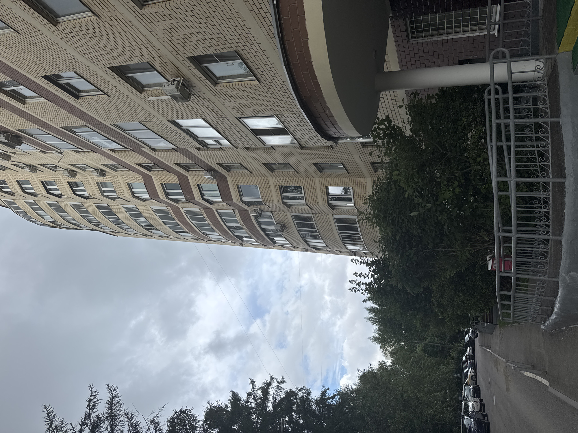 The image shows a view of a light-colored brick apartment building with a curved facade and numerous windows, some of which have air conditioning units installed. The building is in Zelenograd, Russia, as indicated by the context. To the left of the building, there is a street with parked cars and a sidewalk lined with trees and bushes. The sky is overcast with clouds. A metal railing is visible along the sidewalk in the foreground. The scene appears to be an ordinary residential street in a city.
