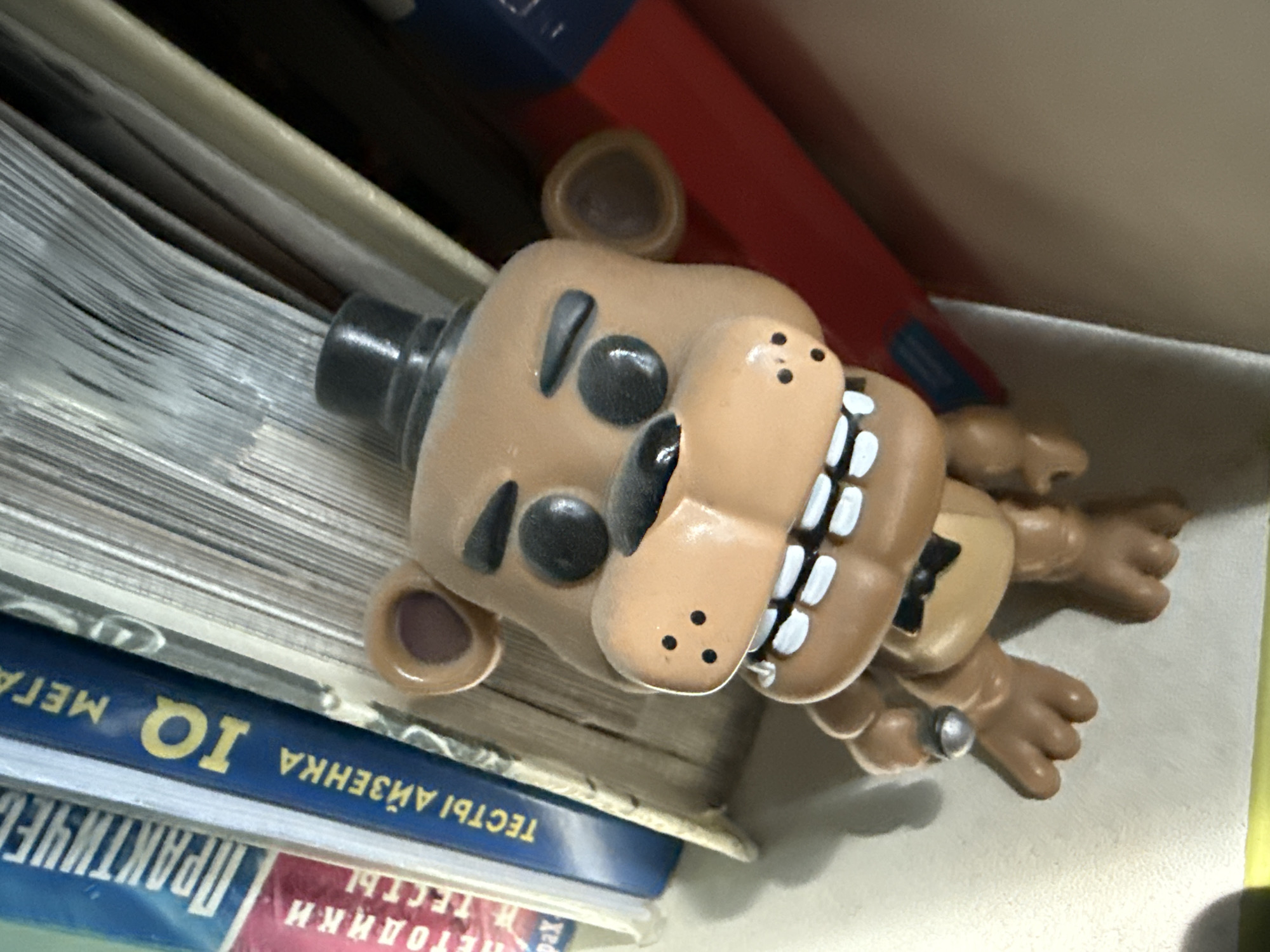 An upright, close-up, slightly angled shot captures a light brown Freddy Fazbear figurine with black eyes, a black top hat, and a black bowtie, positioned between books on a shelf. The figurine's mouth is open, revealing white teeth and a dark interior. Its left hand, holding a small silver object, rests on its chest. Behind the figurine, various books with visible spines are stacked, some displaying Russian text, including "ПРАКТИЧЕС" and "IQ." A predominantly red book is visible to the right, partially obscured. The background is out of focus, suggesting an indoor setting, possibly a home or office. There is no information to determine the location of the city of Simferopol, Ukraine, in this image.