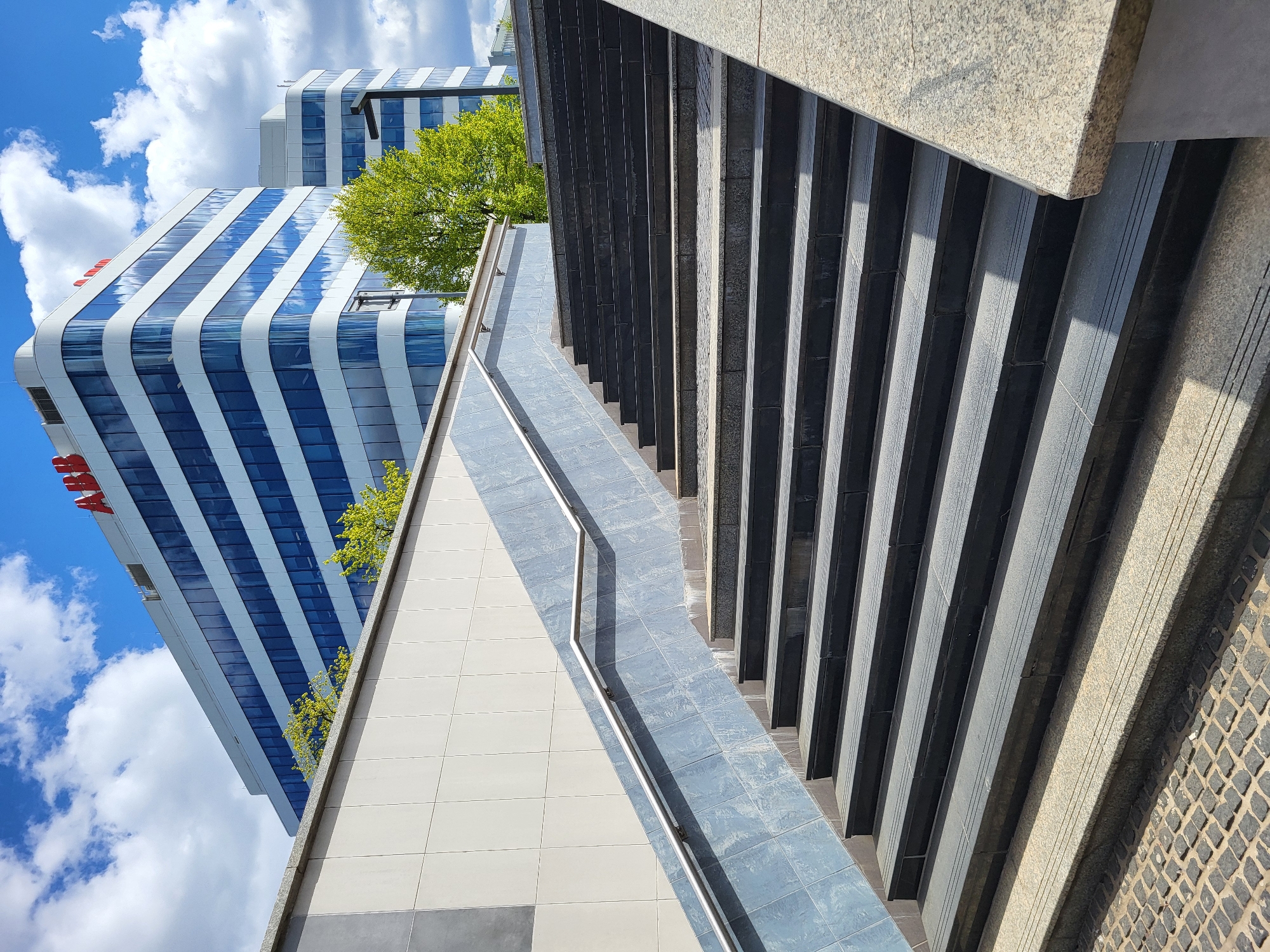 This media file captures an urban architectural scene under bright daylight. The image is rotated 90 degrees counter-clockwise, making the buildings appear on their sides.
The main subjects include two modern buildings with distinctive design, a grand outdoor staircase, a tiled ramp with a handrail, and scattered trees. The sky is bright blue with fluffy white clouds, indicating clear weather.
The primary building on the left (if the image were rotated upright) features a curved facade with prominent horizontal bands of dark blue reflective glass alternating with white or light grey solid strips. On its lower left side, visible in red, is the text "ABB". Another similar but partially obscured building is behind it. Vibrant green foliage from trees is visible in the middle ground, separating the buildings.
In the foreground, a large, wide outdoor staircase dominates the right side of the frame. The stairs are made of dark grey or black stone, with textured surfaces that reflect sunlight, creating visible striations. Adjacent to these stairs, running diagonally through the mid-ground, is a ramp or walkway with a light-colored metal handrail. This ramp is surfaced with light grey and blue-grey tiles, showing a geometric pattern. To the far right, another section of textured stone, possibly part of a wall or another set of stairs, borders an area with dark, cobblestone-like paving.
Strong sunlight casts distinct shadows across the stairs and other surfaces, highlighting their textures and depth. There are no people visible in the scene. The overall impression is one of modern architecture and structured outdoor spaces on a sunny day.