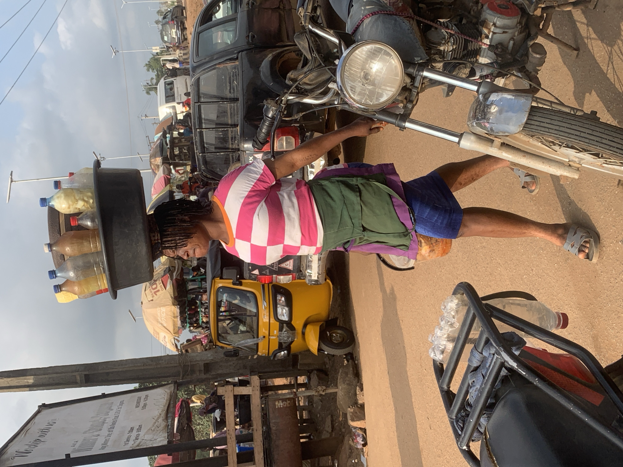 A woman walks with a bowl of bottles balanced on her head. She wears a pink and white striped t-shirt, a green apron, and a blue skirt. In the background, a yellow auto-rickshaw, a black pickup truck, and a motorcycle are visible. The scene appears to be a street in Shagamu, Nigeria, with stalls and people suggesting a market setting.