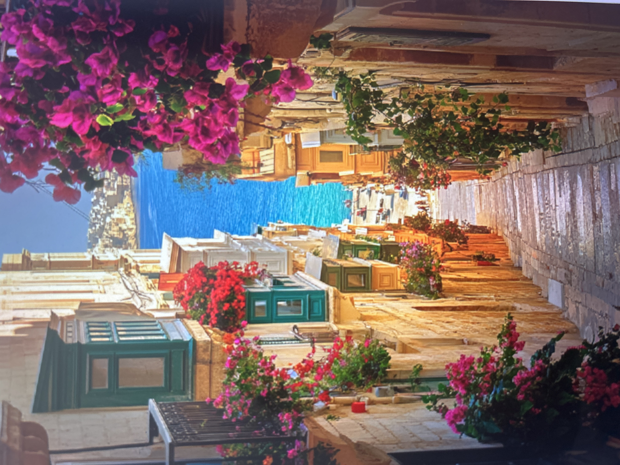 The image depicts a charming, narrow street or alleyway in what appears to be a Mediterranean town. The perspective looks down the street towards a bright blue body of water, likely the sea, in the distance.
The street is lined with traditional stone buildings, many featuring distinctive enclosed wooden balconies, some painted a deep green. These balconies are adorned with an abundance of vibrant pink and red bougainvillea flowers, adding splashes of intense color against the earthy tones of the stone. Other greenery and flowering plants are also visible along the street and on the buildings.
The pathway itself appears to be paved with stone or cobblestones and slopes or steps downwards towards the sea. The bright sunlight suggests a clear daytime, possibly late morning or afternoon, casting a warm glow on the buildings and illuminating the colorful flowers. No people or specific activities are visible, presenting a serene and picturesque street scene. The architecture and dense floral decorations strongly evoke the character of a historic Mediterranean or southern European location. There is no visible text.