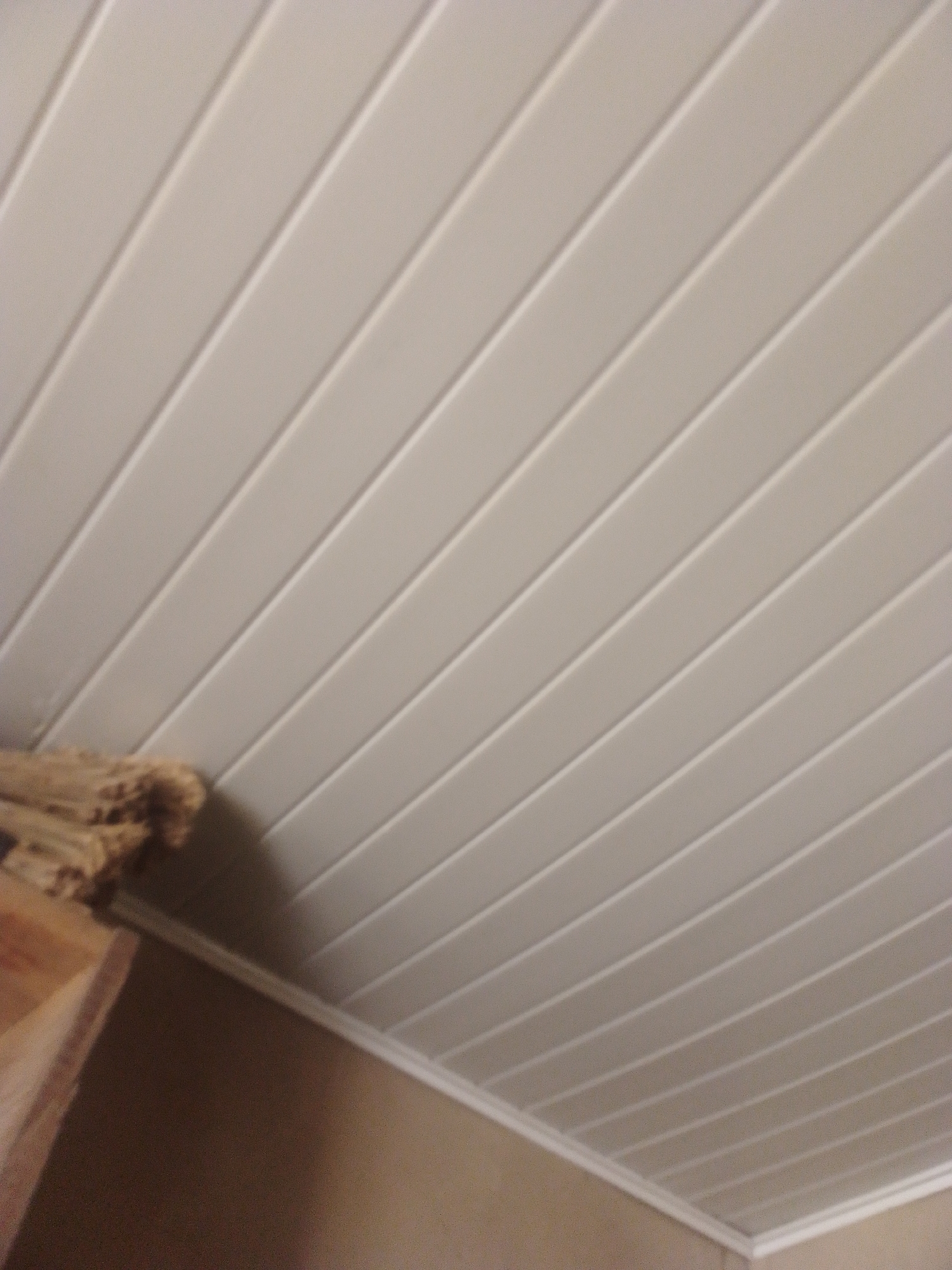 The image shows a close-up view of a paneled ceiling and a portion of a wall. The ceiling is made of light-colored, horizontal panels with visible seams between them, creating a linear pattern. In the lower left corner, there is a stack of what appears to be wood or some other rough material, possibly firewood, resting on a wooden surface. The wall below the ceiling is a plain, muted color, possibly beige or tan. The scene is indoors and well-lit, with the light source likely coming from above and to the right, casting subtle shadows that highlight the texture of the ceiling panels. The image does not provide any specific information about its location within Jauro Yinu, Nigeria.