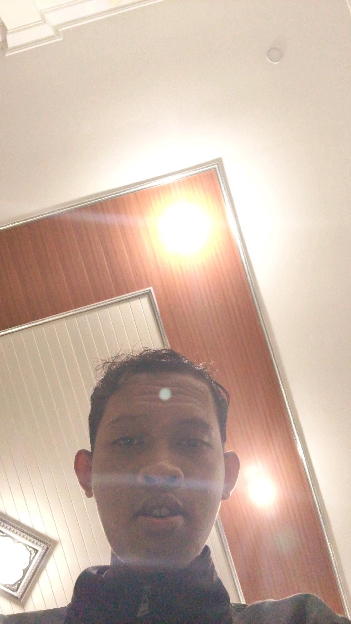 The image is a first-person perspective selfie taken indoors, likely a room with a wood-paneled ceiling. The ceiling features recessed lighting, one of which is prominently visible and bright. The subject's face is partially visible, with a bright light reflection on their forehead. The person appears to be young, with dark hair and an open mouth. To the left of the subject, a framed decorative object is visible, suggesting the setting is a home or an interior space. The overall lighting is dim, with the bright ceiling light causing lens flare. The specific location in the city of Pundungsari, Indonesia cannot be determined from the image alone.