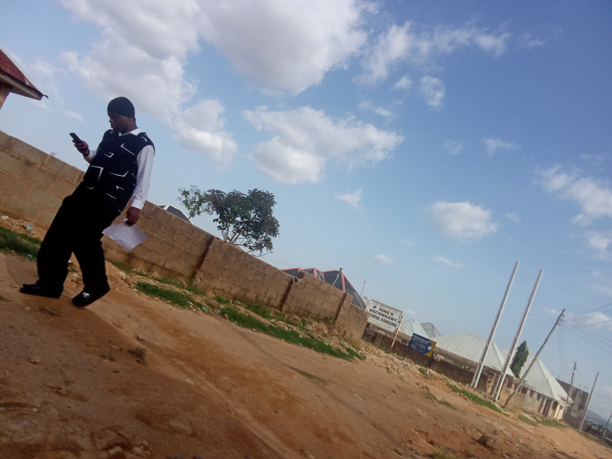 A man in a black vest over a white shirt and black pants walks along a dirt path, holding a cell phone in his left hand and a white paper in his right. He is walking away from the camera, towards a brown brick wall. Behind the wall, a tree with dark green leaves stands tall. In the distance, a sign reads "KING'S SECONDARY SCHOOL KANANG," indicating the location in Barakin Akawo, Nigeria. The sky above is a clear blue, dotted with fluffy white clouds. The ground is a mixture of dirt and sparse green grass.