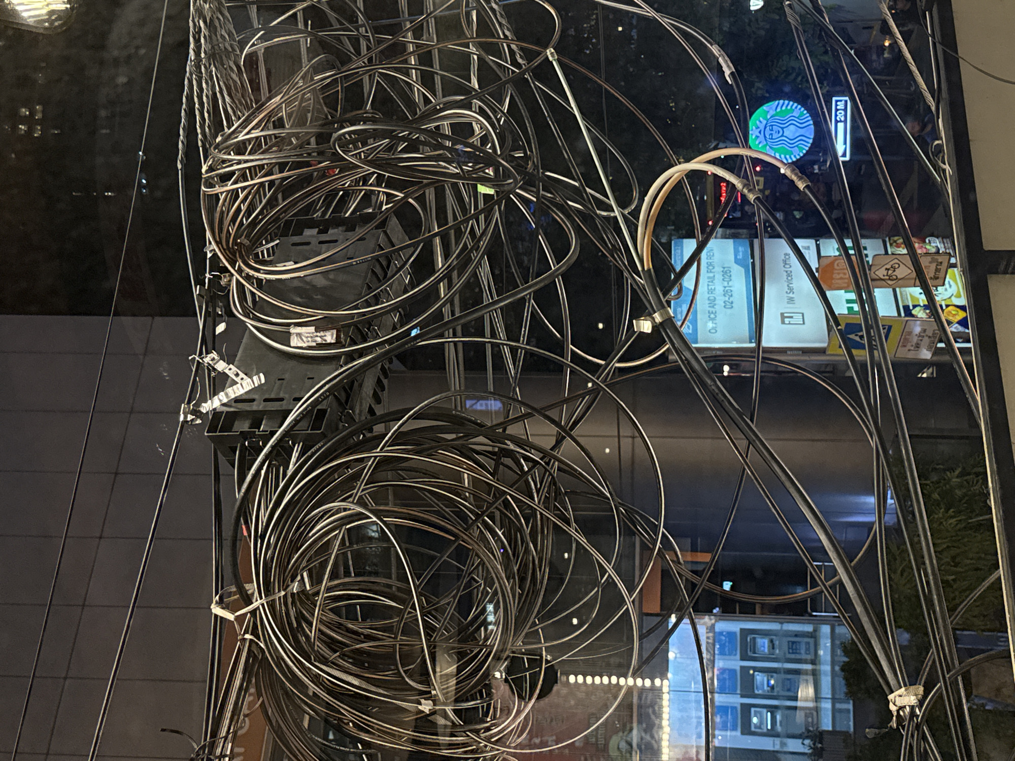 The image captures a dense tangle of black cables and wires, seemingly haphazardly bundled together. These wires are attached to a black, slotted panel on the left side.
Reflections visible in the image provide context. Through the network of wires, there's a glimpse of a Starbucks logo, a street sign indicating "20 M," and other illuminated signs and advertisements in Thai script. One sign visible through the wires promotes "Office and Retail for Rent" with the phone number "02-261-0261." Another sign reads "IW Serviced Office." These details indicate that the location is likely in Bangkok, Thailand, known for its often chaotic overhead cabling infrastructure. The environment appears to be an urban setting at night.