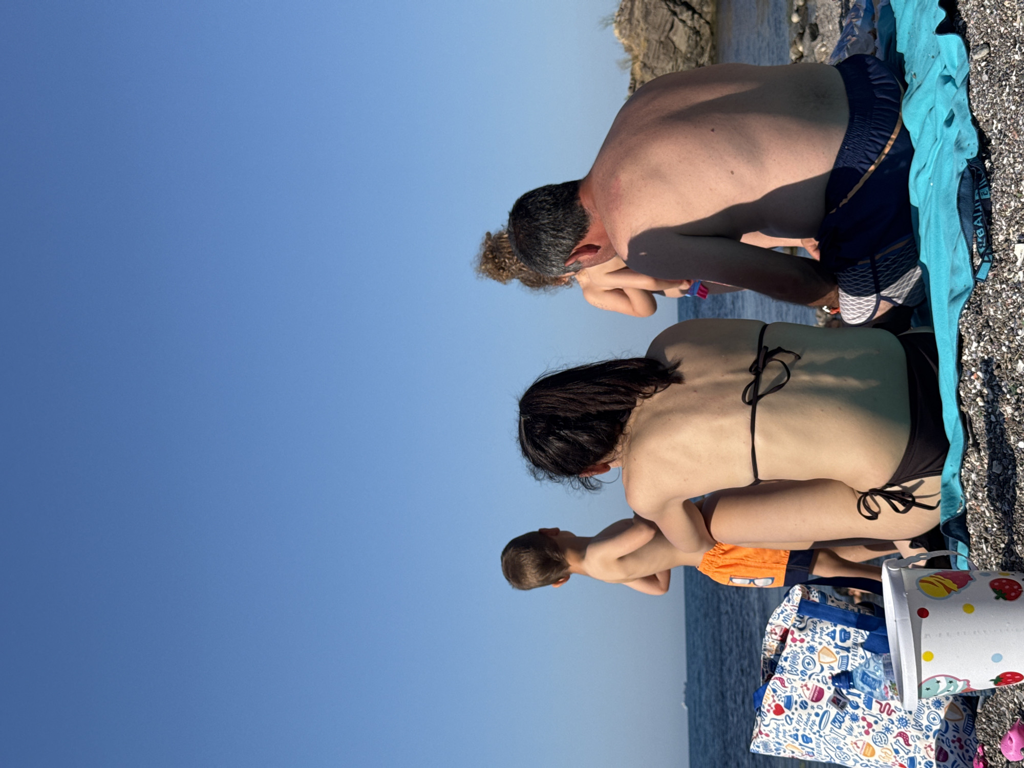 The image captures a family enjoying a sunny day at a beach in Sozopol, Bulgaria. The scene features four individuals, two adults and two children, with a clear blue sky overhead, suggesting fair weather. The adults appear to be a man and a woman, both facing away from the camera, with the man wearing dark blue swimming trunks and the woman in a black bikini. The children are a boy in orange swim trunks, and another child, possibly a younger boy or girl, with curly hair, also appearing to be in swimwear.
The family is positioned on a pebble beach, with a bright blue towel spread out and a partially visible bag with a colorful pattern, possibly a beach bag, and a small white cooler bag with fruit designs. The water of the sea is visible in the background, with gentle waves. The rocky coastline is also present, adding to the natural landscape of the location. The lighting suggests it is likely late afternoon or early evening, indicated by the long shadows cast by the individuals. The overall mood seems relaxed and peaceful, with the family enjoying a typical summer day by the sea. No discernible text is visible in the image.