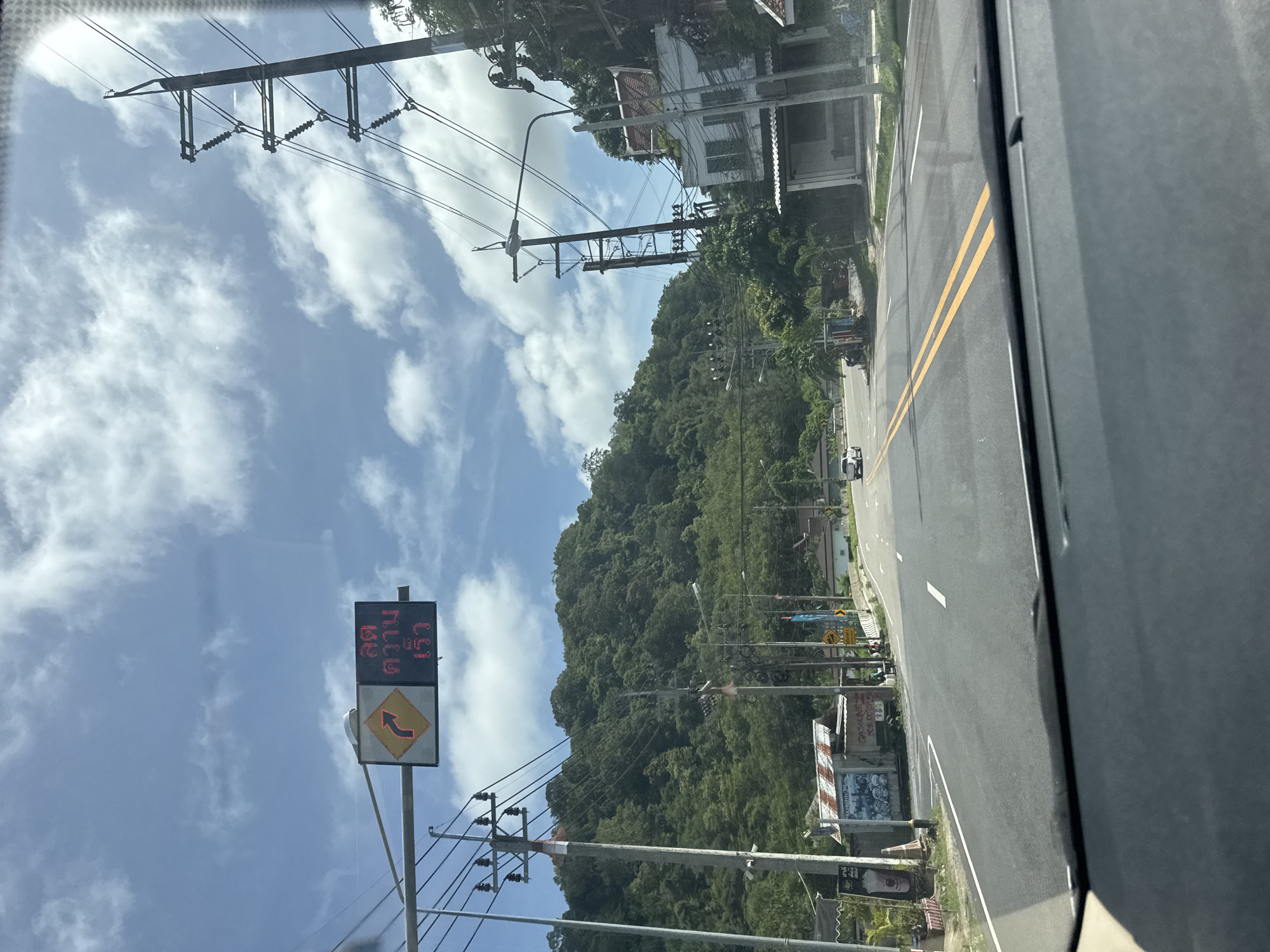 A photograph taken from inside a vehicle looking out at a road in Kamala, Thailand, on a bright, cloudy day. The road is paved with asphalt and has double yellow lines down the center. On the left side of the road, there is a traffic sign with a curved arrow indicating a turn and Thai text in red. Further down the road, a white car is visible. To the right of the road, there are houses and lush green hills in the background. Power lines and utility poles are prominent throughout the scene. The foreground shows the interior of a car, likely the dashboard.