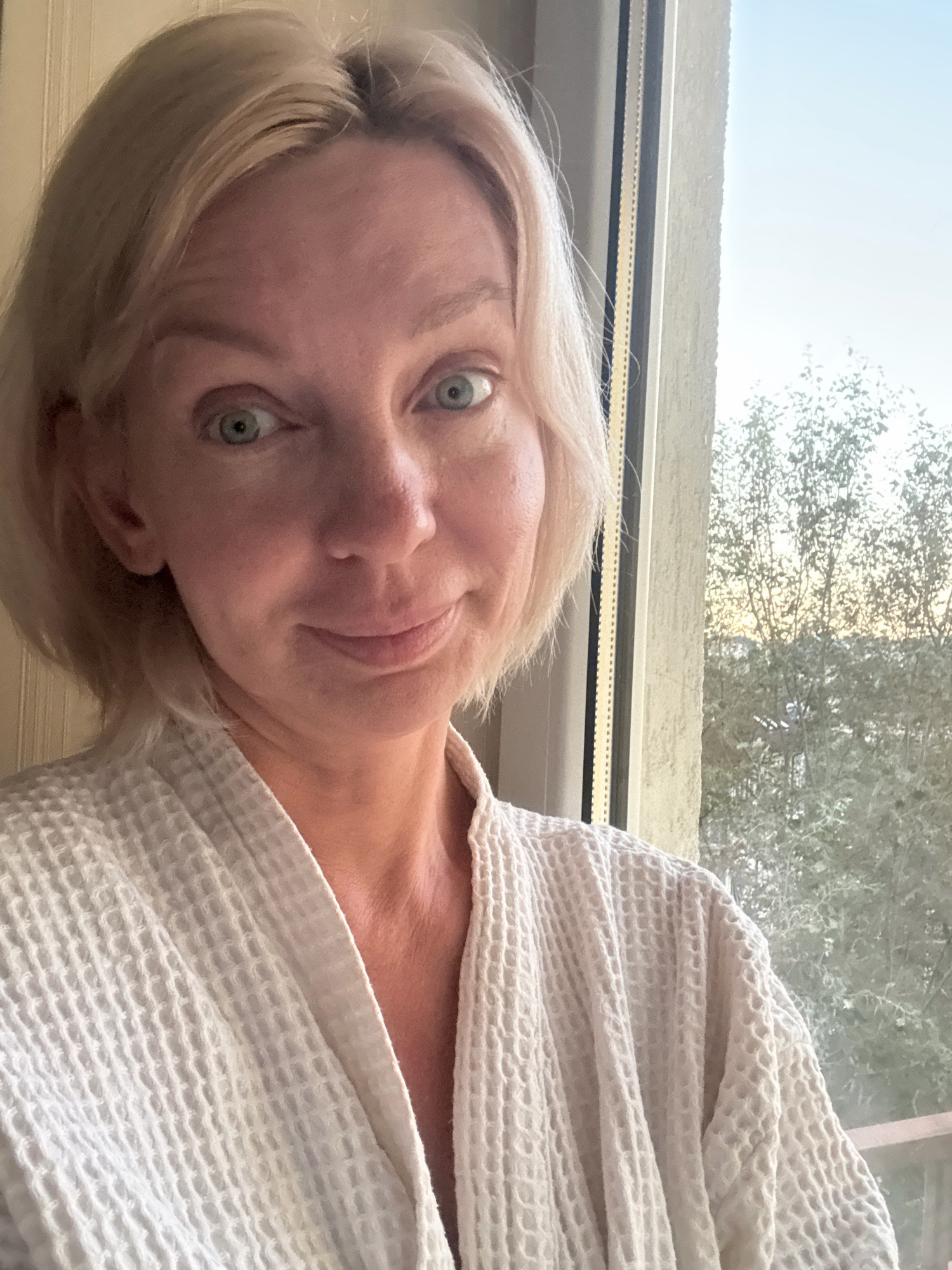 A close-up, head-and-shoulders selfie of a blonde woman wearing a white waffle-weave bathrobe is captured in front of a window. The woman's eyes are wide and looking directly at the camera, with a slight smile. Her blonde hair is cut in a bob. The window behind her offers a view of trees and a bright sky, suggesting an outdoor scene. The lighting suggests it is either early morning or late afternoon. The image does not depict the city of Sochi, Russia.