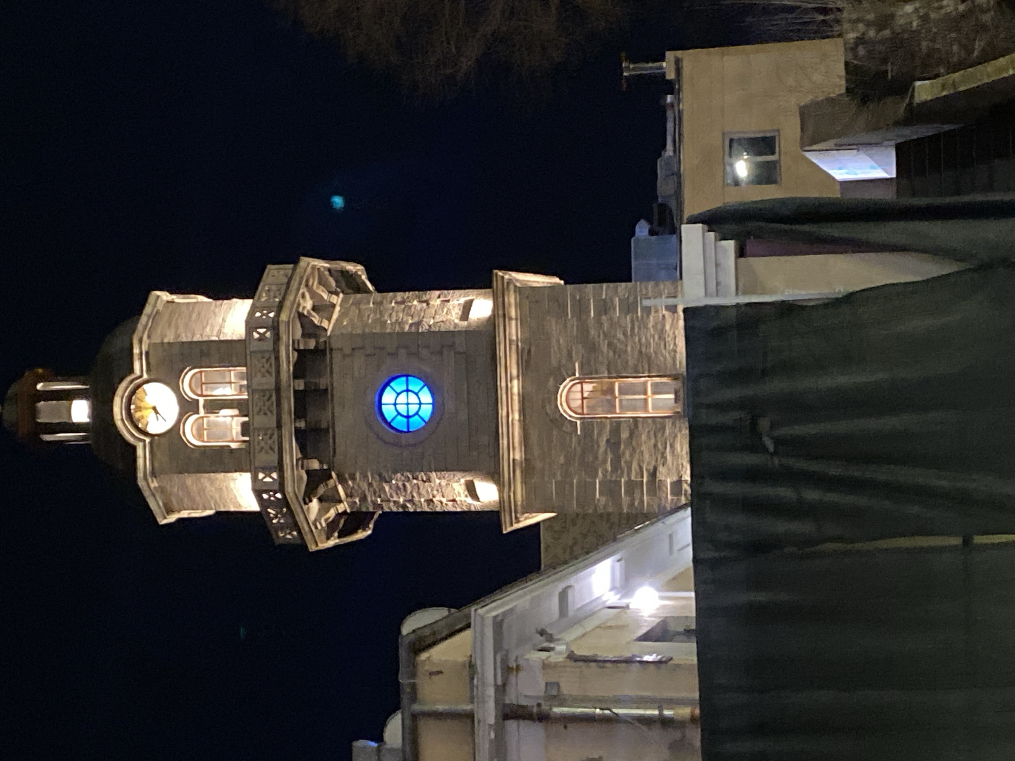 The image shows the clock tower of the Varna Municipality at night. The tower is made of light-colored stone and has a clock face and a blue round window. The tower is illuminated with lights, and the sky is dark. The tower is located in the city center of Varna, Bulgaria. Other buildings are visible surrounding the tower. The photo is taken from an elevated angle. A dark green covering is partially obscuring the view from the right side of the image.