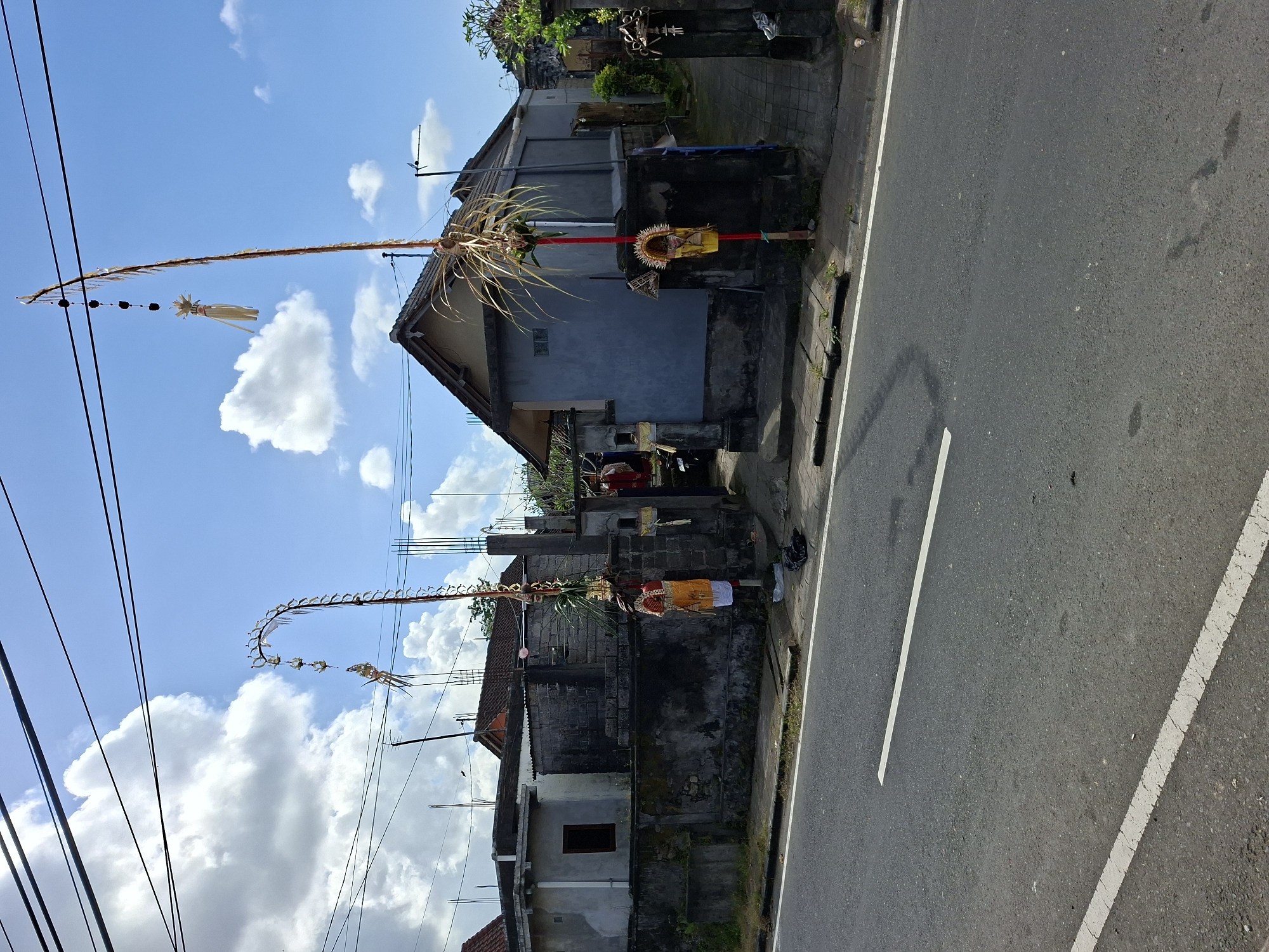 In Blahbatuh, Indonesia, on a bright, partly cloudy day, a street scene unfolds. The foreground is dominated by a paved road with white lane markings, suggesting it is a public thoroughfare. To the left of the road, a sidewalk runs alongside a wall, behind which are residential buildings. These structures are varied in design and condition, with some appearing aged and others under construction.
The most prominent features are two elaborately decorated poles, known as "penjor," which are traditional Balinese offerings. The first penjor, to the left, is tall and arched, adorned with woven palm leaves and small decorative elements. The second penjor, to the right, is a straighter, more slender pole, also decorated with palm leaves and smaller embellishments, including what appears to be a small figure or effigy attached. Both poles are adorned with red and yellow fabrics, indicating a festive or religious occasion.
The background reveals more of the residential area, with houses showing different architectural styles, some with tiled roofs and others with corrugated metal. Power lines crisscross the sky, a common sight in many urban and semi-urban environments. The overall impression is a snapshot of daily life in a Balinese village, marked by cultural traditions and the coexistence of older and newer structures.