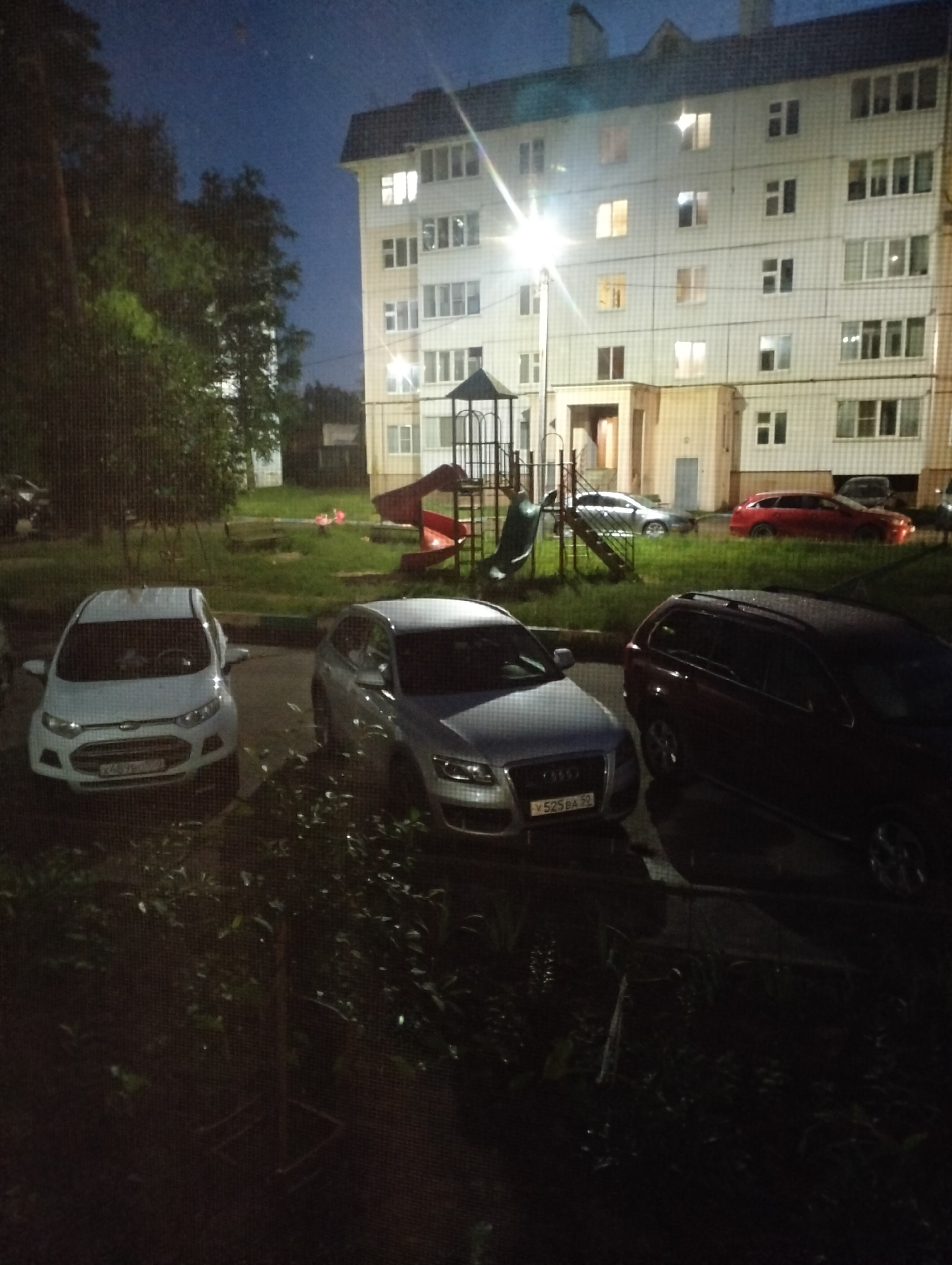 A nighttime view shows a multi-story apartment building with several windows illuminated. In front of the building, a children's playground with a red slide is visible. Parked cars are in the foreground, including a white Ford EcoSport with a license plate reading "Y489BO 52," a silver Audi with the license plate "Y 525 BA 52," and a dark-colored SUV. The scene appears to be a residential area, possibly a courtyard, with trees and grass. The sky is dark, suggesting it is late evening or night. There are no people visible in the image. The overall impression is of a quiet, ordinary residential setting at night.