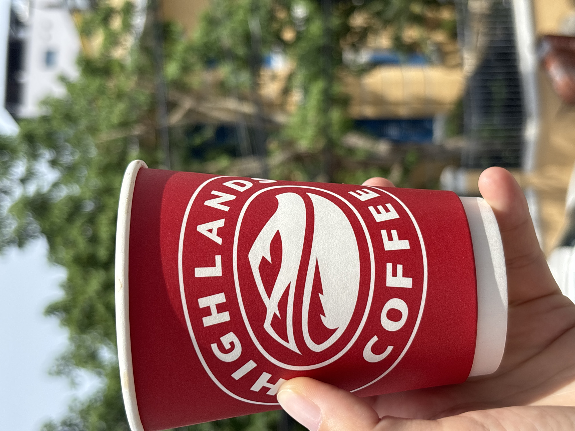 A hand holds a red disposable coffee cup with a white rim, displaying the "Highlands Coffee" logo in white. The logo features a stylized flame or leaf emblem surrounded by text. The cup is tilted, with the bottom portion of the text "HIGHLANDS COFFEE" visible in a circular arrangement. A thumb and index finger are supporting the cup from the bottom. The background is blurred, showing greenery from trees and some indistinct building structures, suggesting an outdoor setting. The lighting indicates it is daytime, likely sunny. The overall scene evokes a casual, on-the-go moment of enjoying a beverage. The location context of Nha Trang Ward, Vietnam, is provided.