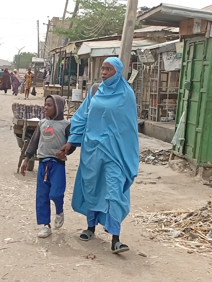 This image depicts a street scene in the city of Gawuna, Nigeria. In the foreground, an adult female and a young male child walk from right to left on a dry, uneven dirt path. The female is dressed in a light blue full-body hijab covering her head and body, light blue trousers, dark socks, and grey slide sandals. Her eyes are visible, and she looks slightly to her right. She carries a dark bag on her left shoulder. The child, walking to her left, wears a grey hooded long-sleeve shirt with a graphic design, blue drawstring pants, and white sneakers. His hood is up, and he holds the woman's left hand.
The path is scattered with debris, including small sticks and refuse, particularly on the right side. To the left of the subjects, a two-wheeled cart carrying stacked cylindrical items is partially visible. The background consists of a row of modest market stalls or shops constructed with corrugated metal roofs and various materials for walls, some appearing weathered. A green metal door or shutter is visible on one structure on the right. Overhead, utility poles and electrical wires are present. Several blurred figures are visible walking further down the street in the distant background.