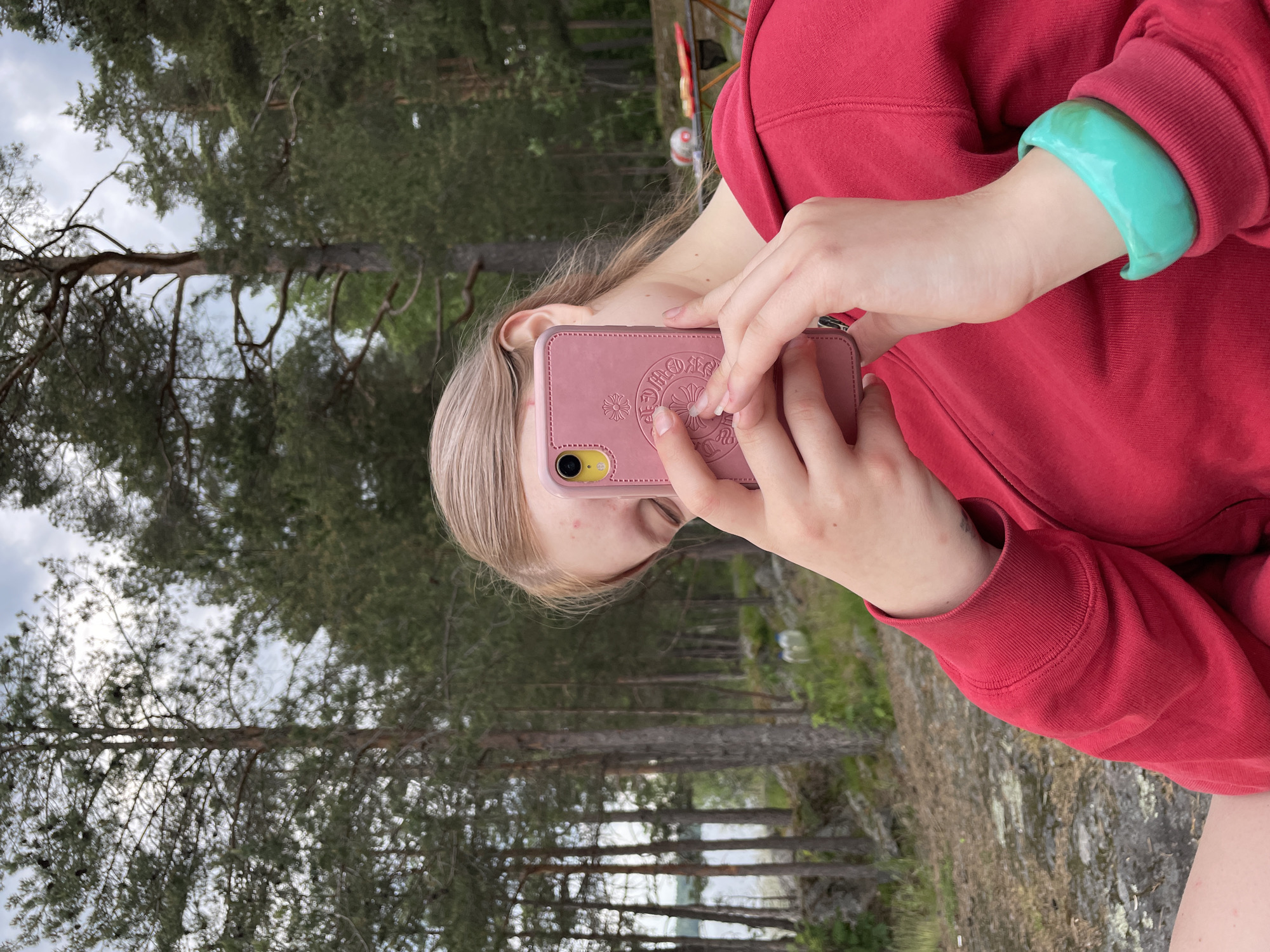 An individual, appearing to be female with blonde hair, is partially visible in an outdoor setting in Yarkoe, Russia. She is wearing a red hooded sweatshirt and a turquoise-colored bangle on her right wrist. Both hands hold a pink smartphone, which obscures most of her face. The phone case features an embossed emblem resembling a floral or cross pattern, and the visible camera module has a yellow outline. The immediate ground is rocky and covered with moss. The background consists of numerous pine trees with dark green foliage. Through the trees on the left, a calm body of water is visible. In the right background, a brown folding table with a red tray and other objects is present. The sky is overcast.