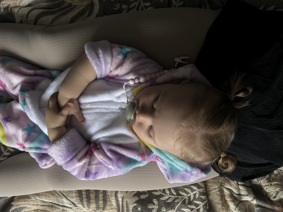 A young child, approximately a toddler, is depicted sleeping on their back, eyes closed, with a green pacifier in their mouth. The child wears a multi-colored plush hooded sleeper patterned with stars, primarily in purple, pink, blue, and yellow hues, over a white undergarment. A pink and white beaded pacifier clip with a small pink figure is attached to the pacifier and draped over the child's chest. The child's light-colored hair is parted down the middle and styled into two small pigtails secured with elastics. The child rests on the light beige, ribbed fabric of an adult's legs or lap. To the left, a patterned fabric, possibly upholstery or a blanket, featuring light floral or leafy motifs on a darker background, is visible. A dark, crumpled fabric is beneath the child's head. The image does not contain any visual elements that identify a specific geographical location, including the city of Leninakan, Russia.