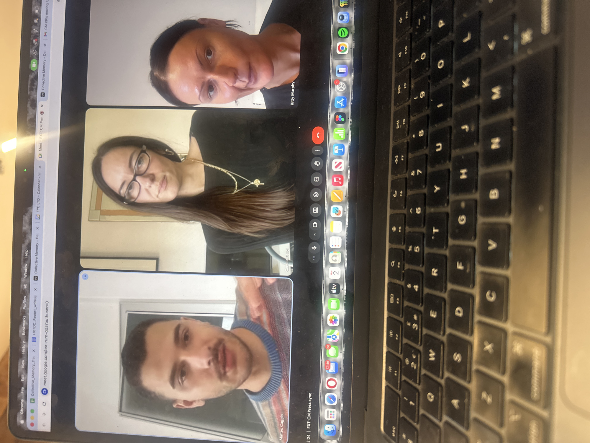 The image depicts a close-up view of a dark grey Apple MacBook Pro laptop screen and part of its keyboard, indicating the scene is located in the city of London, United Kingdom. The screen displays a Google Chrome browser window open to a Google Meet video conference, along with multiple other tabs, including "Collective_Memory_Trac," "HKTDC_Report_writeup," and "EXT: CM Press sync" for the current meeting.
Three individuals are visible in the video conference:
1. **Matteo Caggia** (left panel): A male with dark hair and a mustache, wearing a multi-colored striped sweater with a blue collar. He is looking forward, with his mouth slightly ajar, suggesting he is speaking or listening. His background is a light-colored wall with a white door frame.
2. **Camilla Felici** (center panel): A female with long dark hair, wearing glasses, a black top, and two gold necklaces, one with a key pendant. She is looking directly at the camera with a neutral expression. Her background is a light wall with a framed item on the left.
3. **Kitty Murphy** (right panel): A female with dark hair, wearing a black top. She has a neutral expression with her mouth slightly open. Her background is a plain, light-colored wall.
Below the video conference, the macOS dock is visible, displaying numerous application icons, including Mail (with 17,189 unread messages) and Messages (with 80 unread messages). The physical keyboard, partially visible in the foreground, features black keys with white English QWERTY lettering and shows minor dust accumulation. The laptop's trackpad is also partially visible. The primary action is a virtual meeting in progress, with participants engaged in a collective interaction via the digital platform. The environment is consistent with a personal workspace.