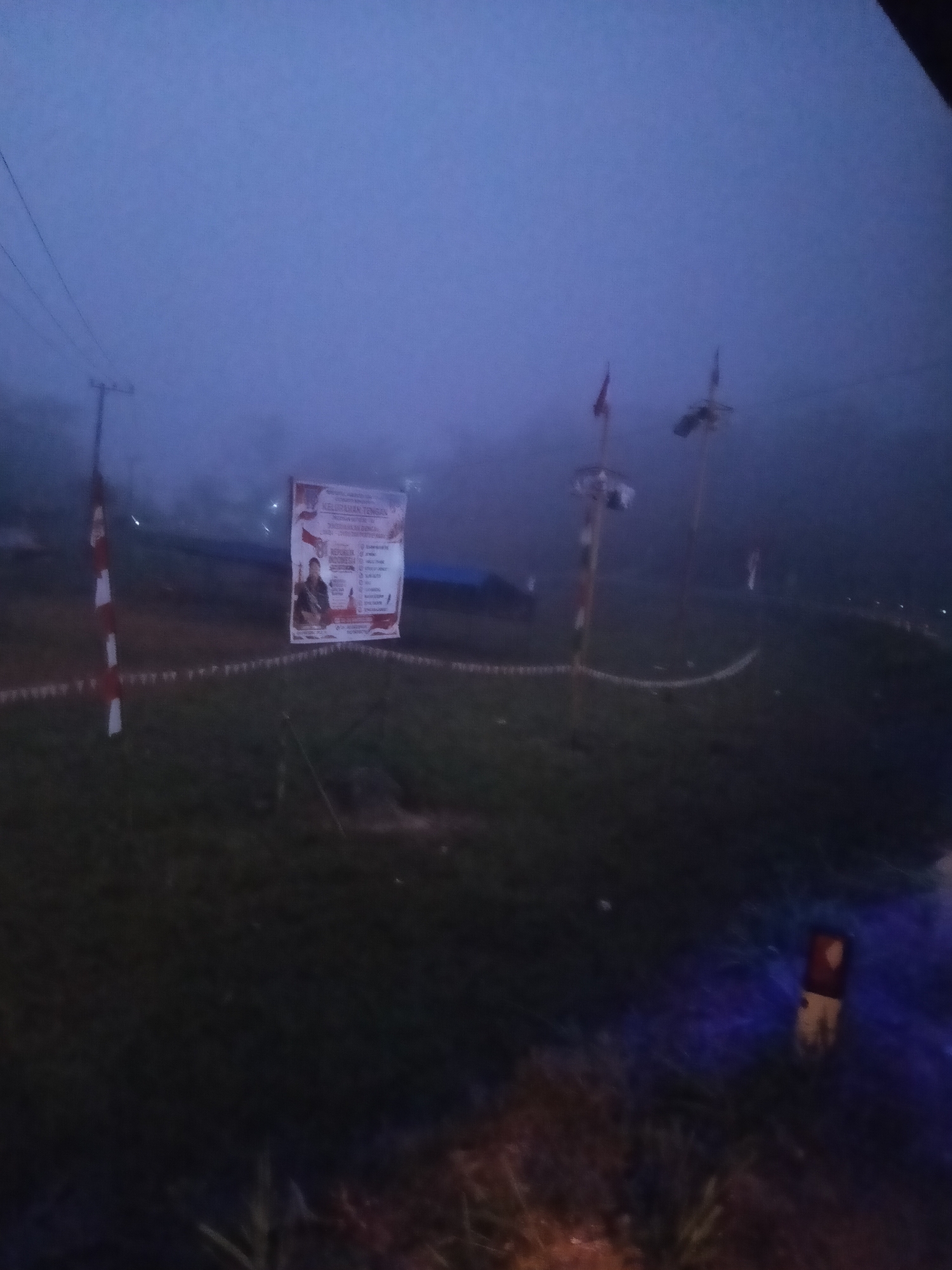 The image captures a foggy outdoor scene at what appears to be dawn or dusk in Tengan, Indonesia. A white and red striped pole stands to the left, possibly marking a boundary or entrance. In the mid-ground, a banner is displayed, featuring images of two individuals and text in Indonesian. The banner is supported by poles, and other poles with small flags are visible further into the background, some obscured by fog. The ground is covered in grass. To the far right, a red and tan vertical object is illuminated by a blue light. The overall atmosphere is hazy due to the thick fog, which significantly reduces visibility.