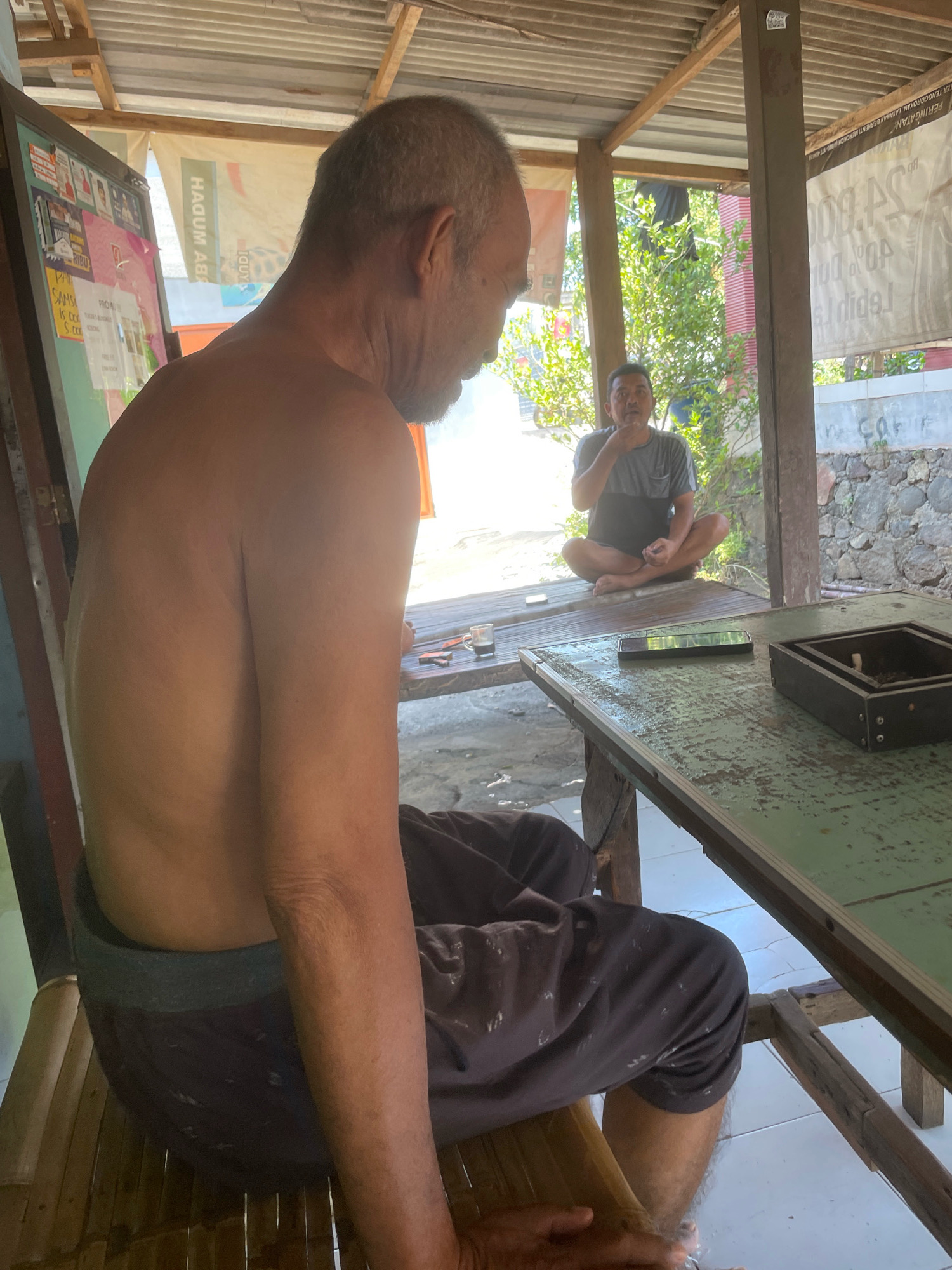 In an outdoor, open-air structure with a corrugated metal roof supported by wooden beams, two men are present. The primary subject, an older man, is seated on a bamboo chair, facing away from the camera, with his back and shoulder exposed. He is wearing dark, loose-fitting pants. To the right, a second man is seated cross-legged on a wooden platform, facing the camera, wearing a grey t-shirt and dark shorts. He is looking off to the side with his hand near his chin. In the foreground, a wooden table with a faded green surface is visible. On the table, there is a mobile phone, a glass with a dark liquid, and a dark rectangular object, possibly an ashtray. In the background, there are trees and stone walls, suggesting a semi-rural or urban fringe location. A banner with Indonesian text is visible on a structure behind the seated men. The lighting indicates it is daytime. The scene is likely in Kebon Waris, Indonesia, given the text in the image.