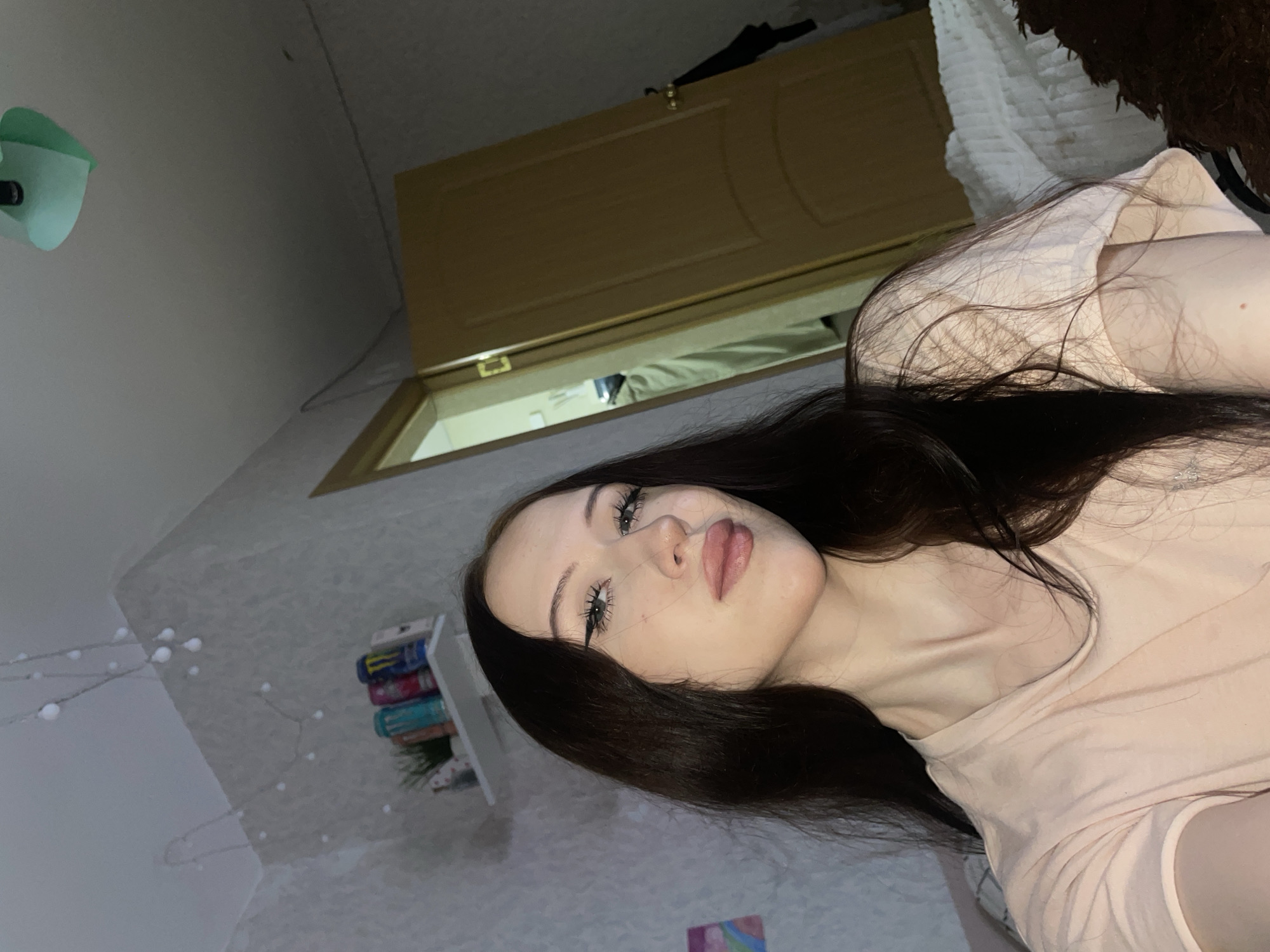 A young woman with dark hair and light skin is lying on a bed in a bedroom. She is wearing a light pink t-shirt. Her head is slightly tilted, and she is looking towards the camera. She has on winged eyeliner. To her left, a shelf is mounted on the wall, holding a small potted plant and several cans of energy drinks. String lights are draped across the upper part of the wall. A closed wooden door is visible in the background, slightly ajar to reveal a glimpse of another room. A portion of a white, ruffled bedspread is on the right side of the frame. The room appears dimly lit, with a soft glow emanating from the string lights. The scene is consistent with an indoor setting in Votkinsk, Russia.