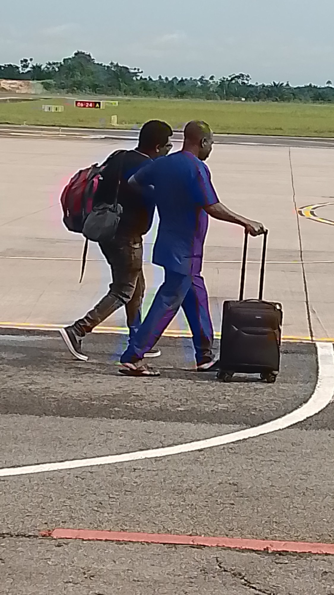 Two men are walking across an asphalt surface, possibly an airport tarmac, with a backdrop of lush green trees under a cloudy sky. The man in the foreground is wearing a blue dashiki and pants, and is pulling a black rolling suitcase. He has dark skin and a shaved head. The man behind him is wearing a black shirt and dark pants, and has a red backpack slung over his right shoulder. He is looking down at his feet. The scene appears to be an airport due to the markings on the ground, including white lines and a red strip. In the background, there are yellow signs with black text, one of which reads "06-24 A". The lighting suggests it is daytime. The overall mood is neutral, with no strong emotional cues. The activity is travel, as indicated by the luggage and the airport setting.