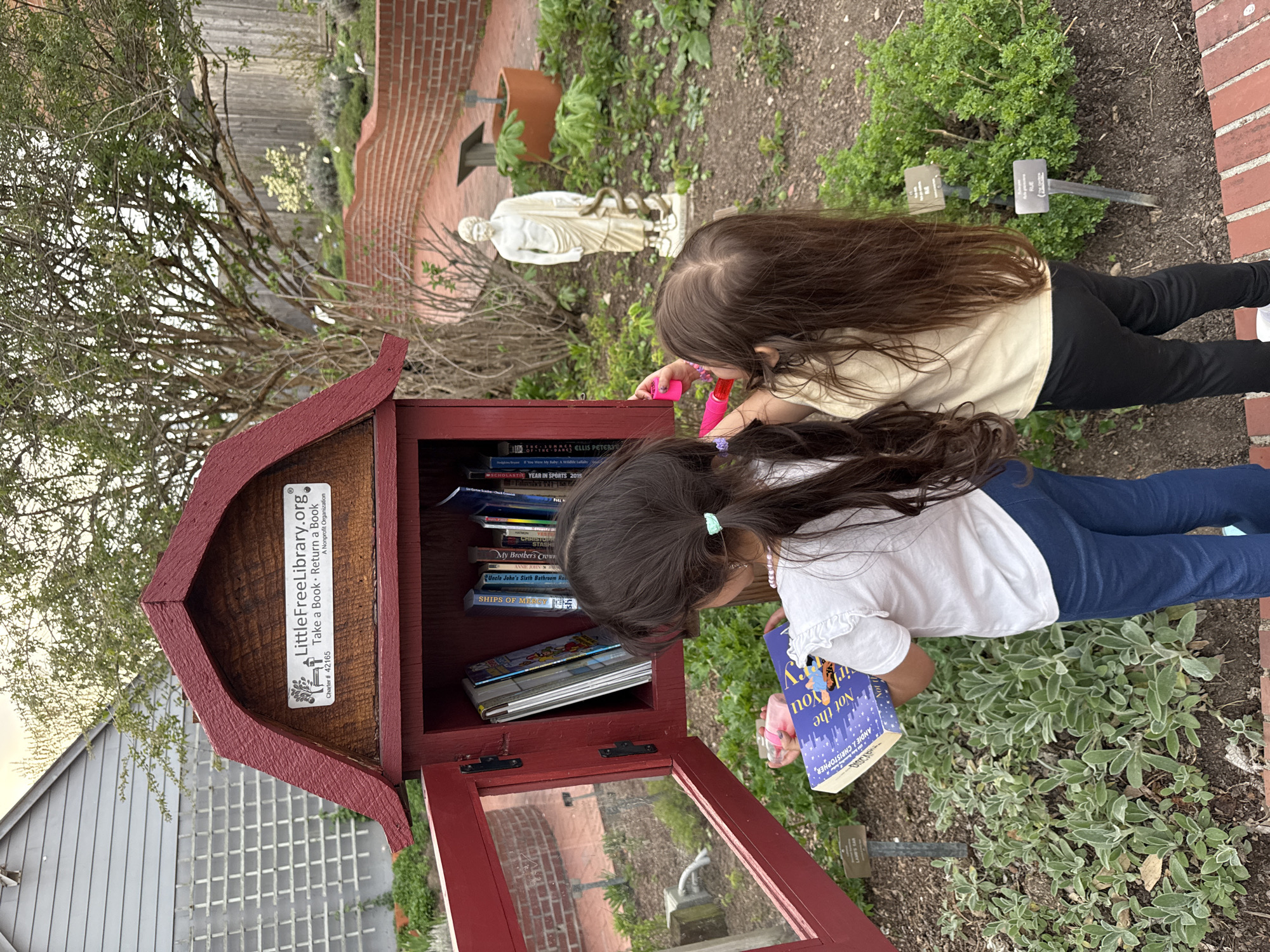 Two young girls are browsing a Little Free Library. The library is a small, red wooden structure with a glass door and is located in an outdoor setting with gardens, brick pathways, and a statue. The library's sign reads "LittleFreeLibrary.org, Take a Book Return a Book, Charter # 42165, A Nonprofit Organization". One girl is holding a book titled "Not the You" by Andie J. Christopher. The books inside the library include "THE SUMMER OF THE DARKS ELLIS PETERS", "SCHOLASTIC YEAR IN SPORTS 2015", "My Brother's Crown", "ANNIE JOHN", "Uncle John's Sixth Bathroom Res", and "SHIPS OF MERCY". The location is in Grove City, United States of America.