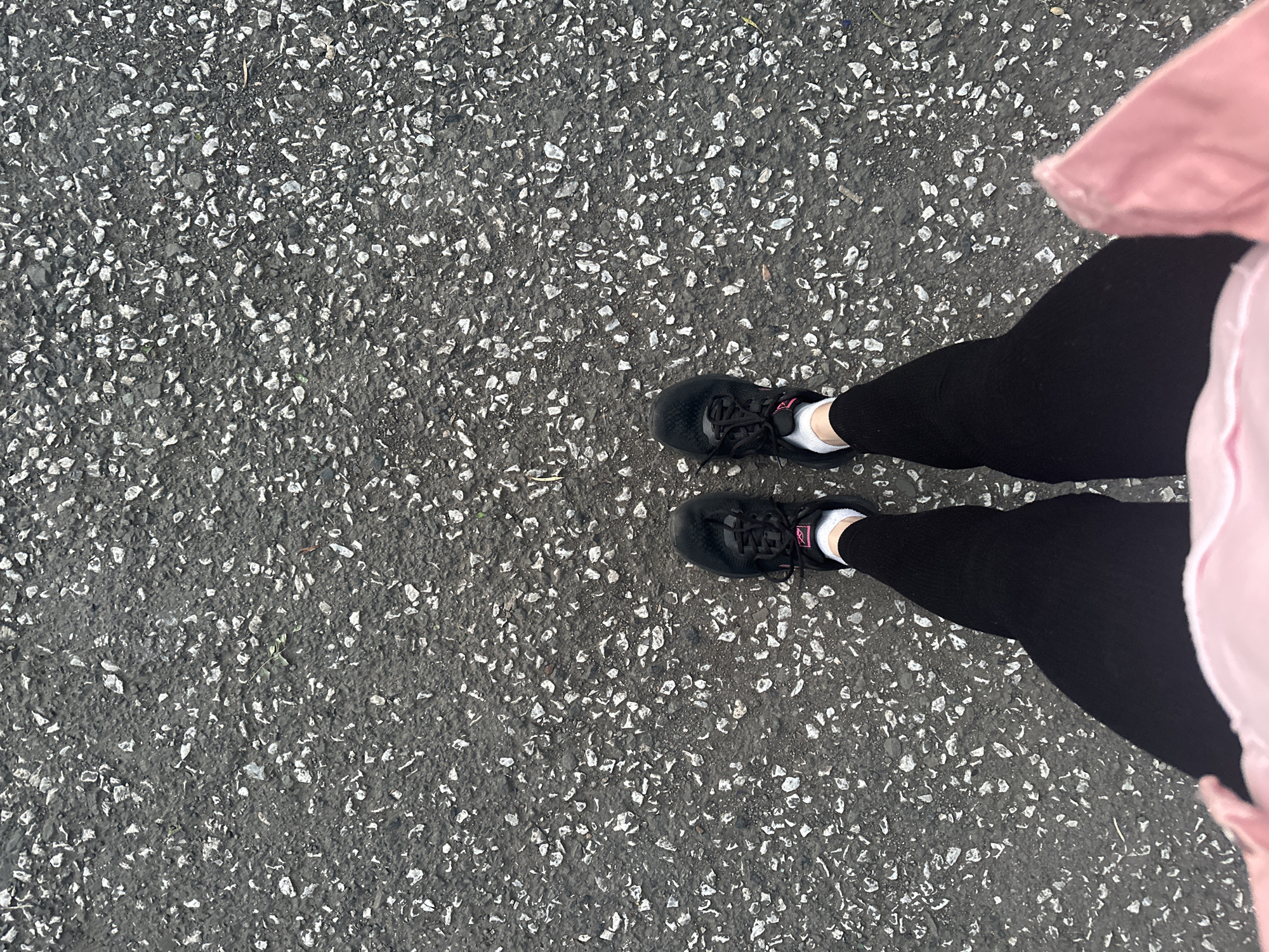 The image shows a top-down view of a person's legs and feet standing on asphalt in Magnitogorsk, Russia. The person is wearing black athletic shoes with pink accents, white socks, and black leggings. A pink jacket is visible at the bottom of the frame. The asphalt is dark gray and textured with small, light-colored stones. There are no other people or objects clearly visible in the scene. The lighting suggests it is daytime.