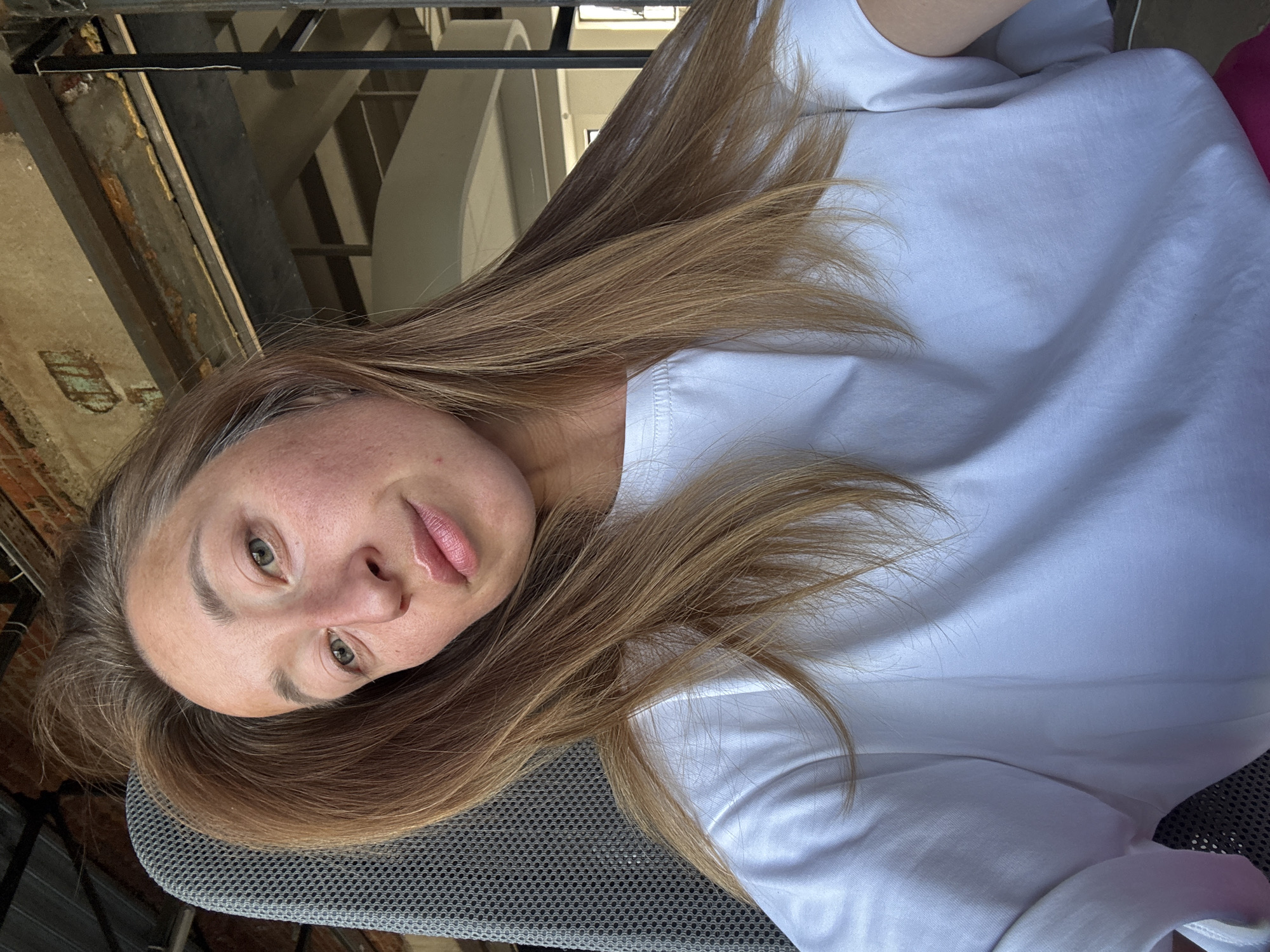 A woman with long, light brown hair is looking at the camera. She is wearing a white t-shirt and appears to be seated on a grey chair. The background includes what looks like a building structure and possible stairway. The location is Chaikovsky, Russia.