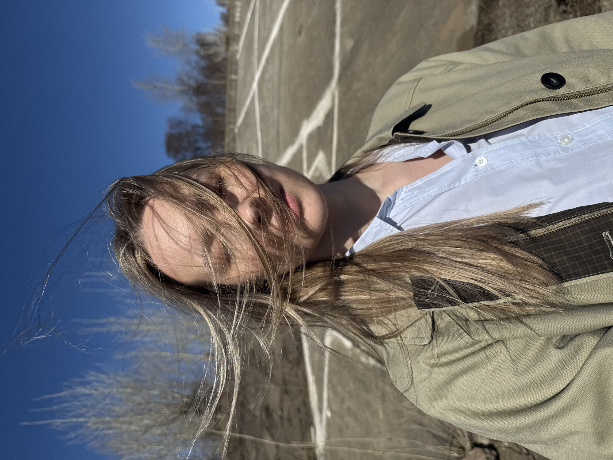 A photograph shows a woman in Chaikovsky, Russia. The woman has long, light brown hair and is wearing a light blue button-down shirt under a light green jacket. The jacket has a single black button visible near her shoulder. The photograph is taken from a low angle, and the woman's face is partially obscured by her hair. In the background, there is a clear blue sky and some bare trees. The ground appears to be concrete with white lines painted on it, possibly a parking lot or a similar paved area. The light suggests it is a sunny day.