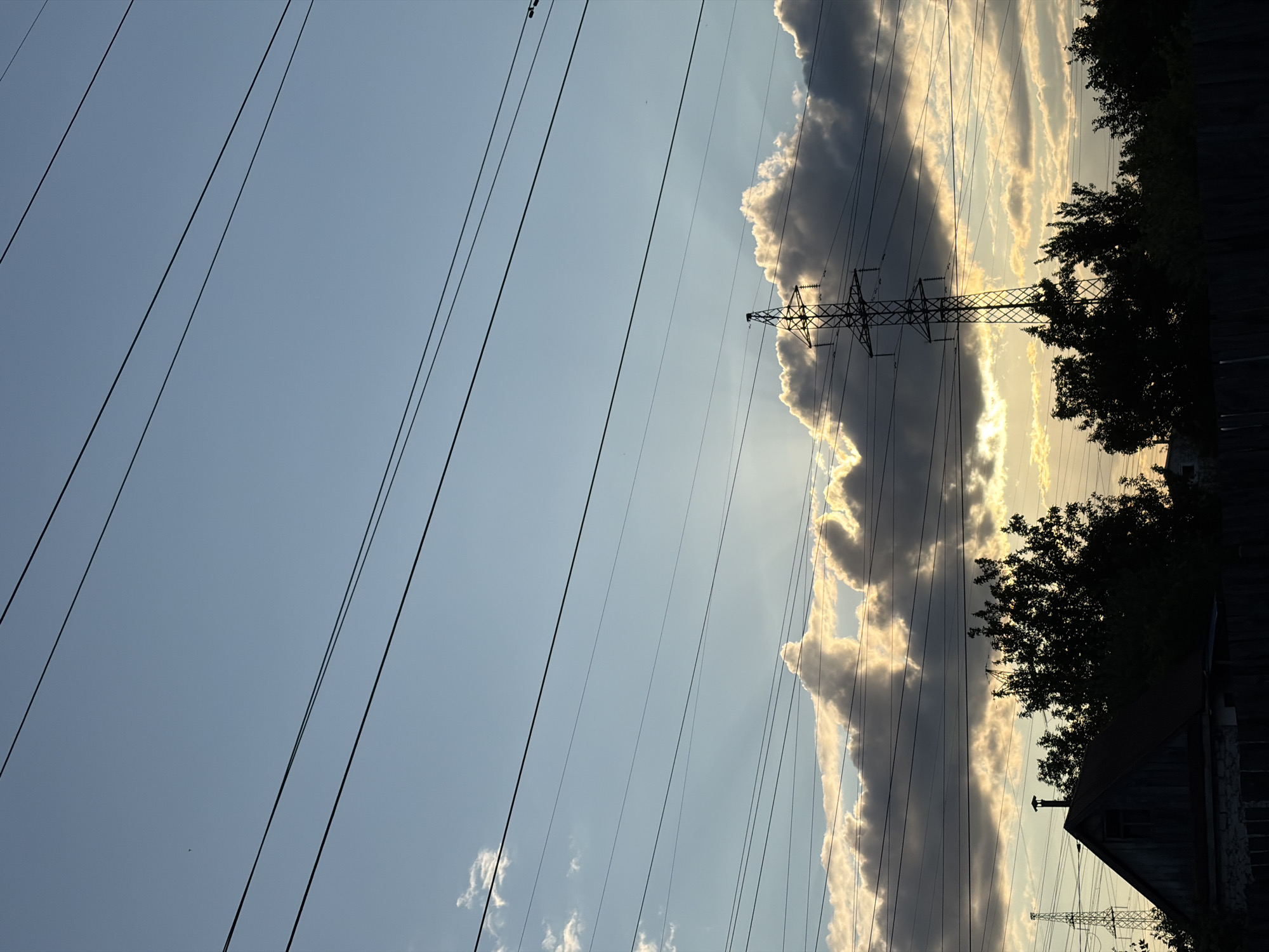 An outdoor, eye-level, medium shot captures a sunset scene in Chaikovsky, Russia. The sky is a gradient of pale blue and grey with scattered clouds. Several power lines stretch across the frame. In the lower portion of the image, silhouetted against the sky, are trees, a small house with a pitched roof, and a large power transmission tower. The sun's rays are visible through gaps in the clouds, creating a dramatic effect. The overall mood is peaceful and serene, with a hint of industrial presence from the power lines and tower.