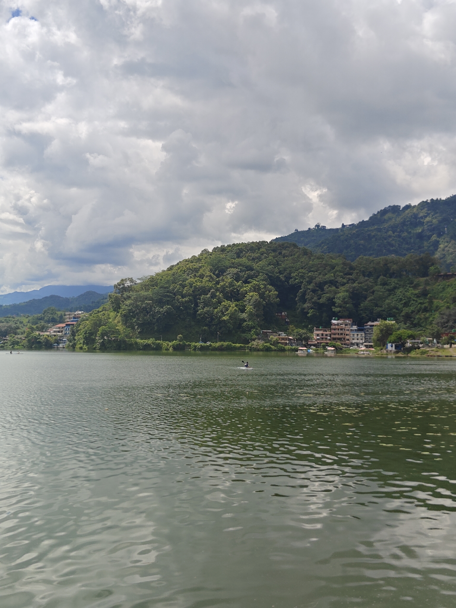 This is a photograph taken from the waters of Phewa Lake in Pokhara, Nepal, looking towards the shoreline. The sky is filled with dramatic, grey clouds, suggesting an overcast or impending weather change. The foreground is dominated by the rippling, dark green water of the lake.
In the middle ground, the tree-covered hills rise steeply from the water's edge. A collection of buildings, appearing to be a mix of residential and commercial structures, are nestled among the trees on the hillside. These buildings are of varying heights and colors, with some featuring balconies and multi-story designs. Several small boats are visible near the shore.
Further out on the lake, two individuals are engaged in recreational activity, appearing to be paddling a small boat or kayak. In the distance, more rolling hills and mountains are visible, partially obscured by the clouds and haze. The overall scene depicts a natural landscape with signs of human habitation and activity along the lake.
