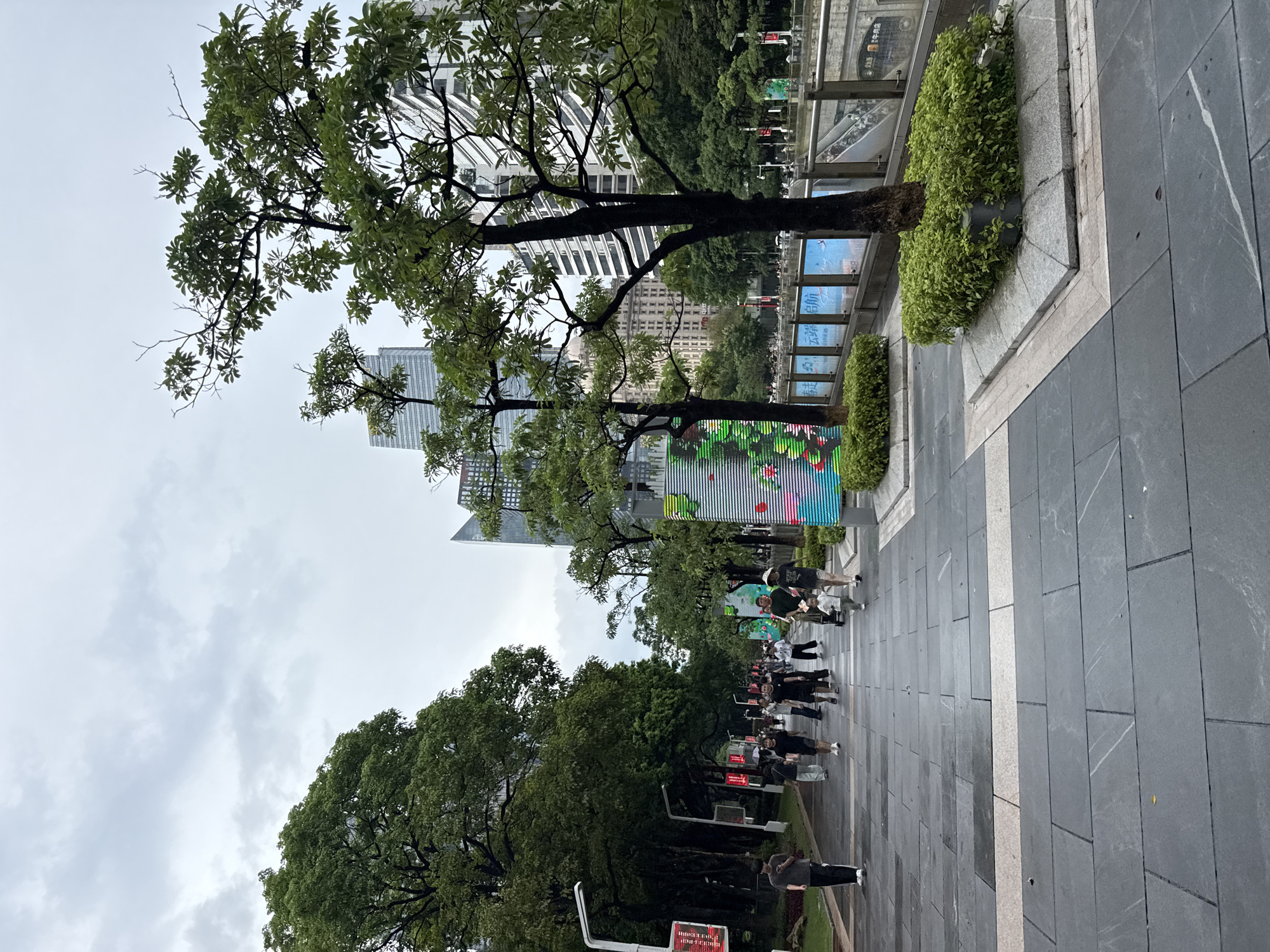This image depicts a pedestrian walkway in Tianhe District, China, on an overcast day. The walkway is paved with large, dark gray rectangular stones, with a lighter colored stone stripe running down the center. Lush green trees line the left side of the walkway, casting shadows. On the right, a canal or waterway is visible, bordered by a metal railing. Beyond the railing, there are escalators leading down. In the background, modern glass and steel buildings rise against the cloudy sky. Several people are walking along the pathway. Some are dressed in casual attire, while others appear to be pushing strollers. There are also decorative elements on the right side of the walkway, including illuminated panels with green and colorful designs, and planters with manicured green bushes surrounding the bases of trees. A street lamp with a red digital display is visible on the left side of the walkway. The ground appears damp, suggesting recent rain.