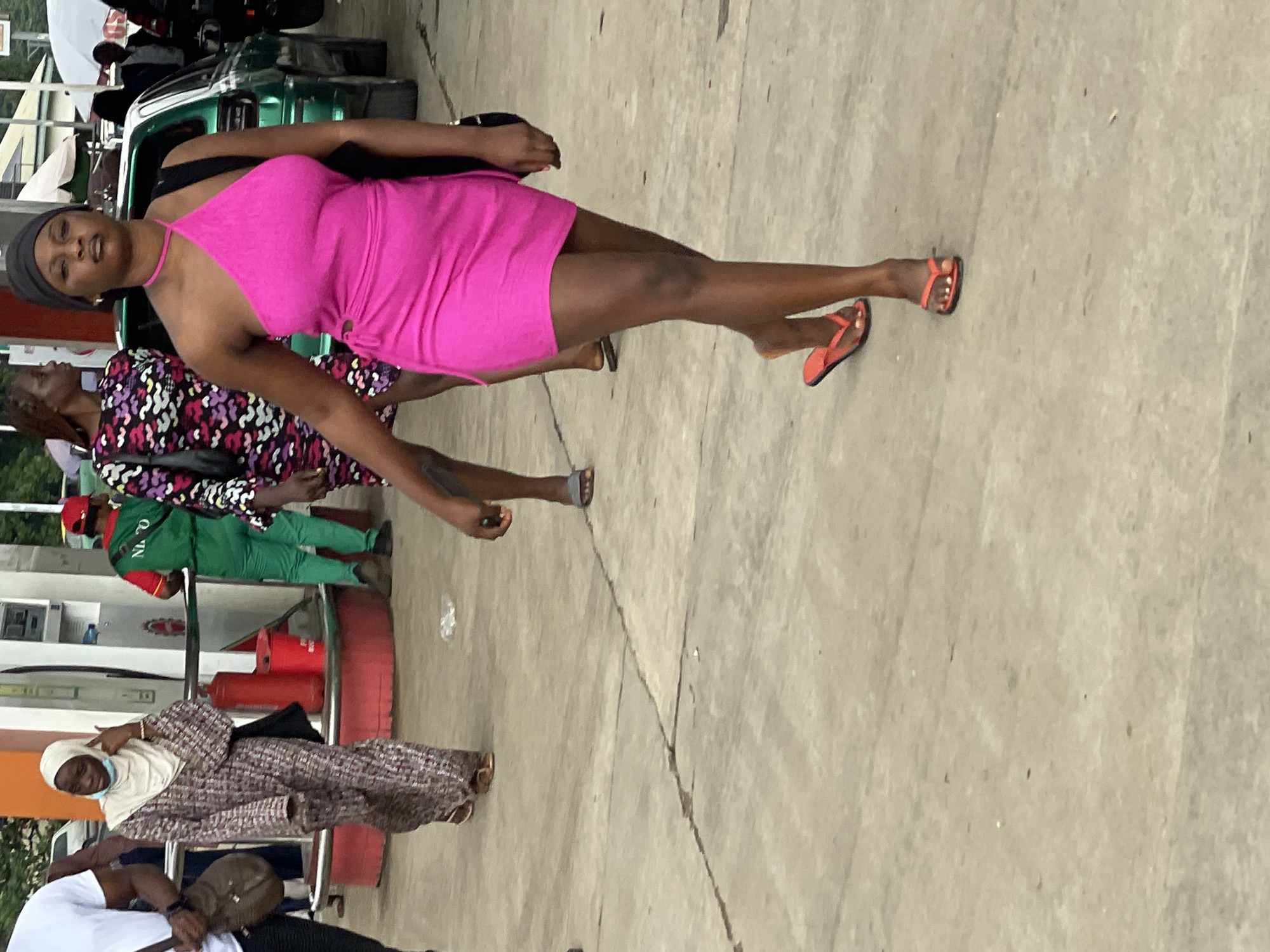 The image shows a street scene in Abuja, Nigeria, likely near a gas station, indicated by the presence of fuel pumps and a green vehicle. In the foreground, a woman in a bright pink, one-shoulder dress with cut-out detailing walks towards the viewer. She wears a black headscarf and orange flip-flops. Behind her, another woman in a patterned dress and a man in a green uniform with "NLPO" on the back are also visible. To the left, a woman in a patterned abaya and headscarf walks away from the camera, carrying a brown bag. The ground is paved concrete, and there are visible cracks and debris. The overall atmosphere suggests a busy, everyday setting.