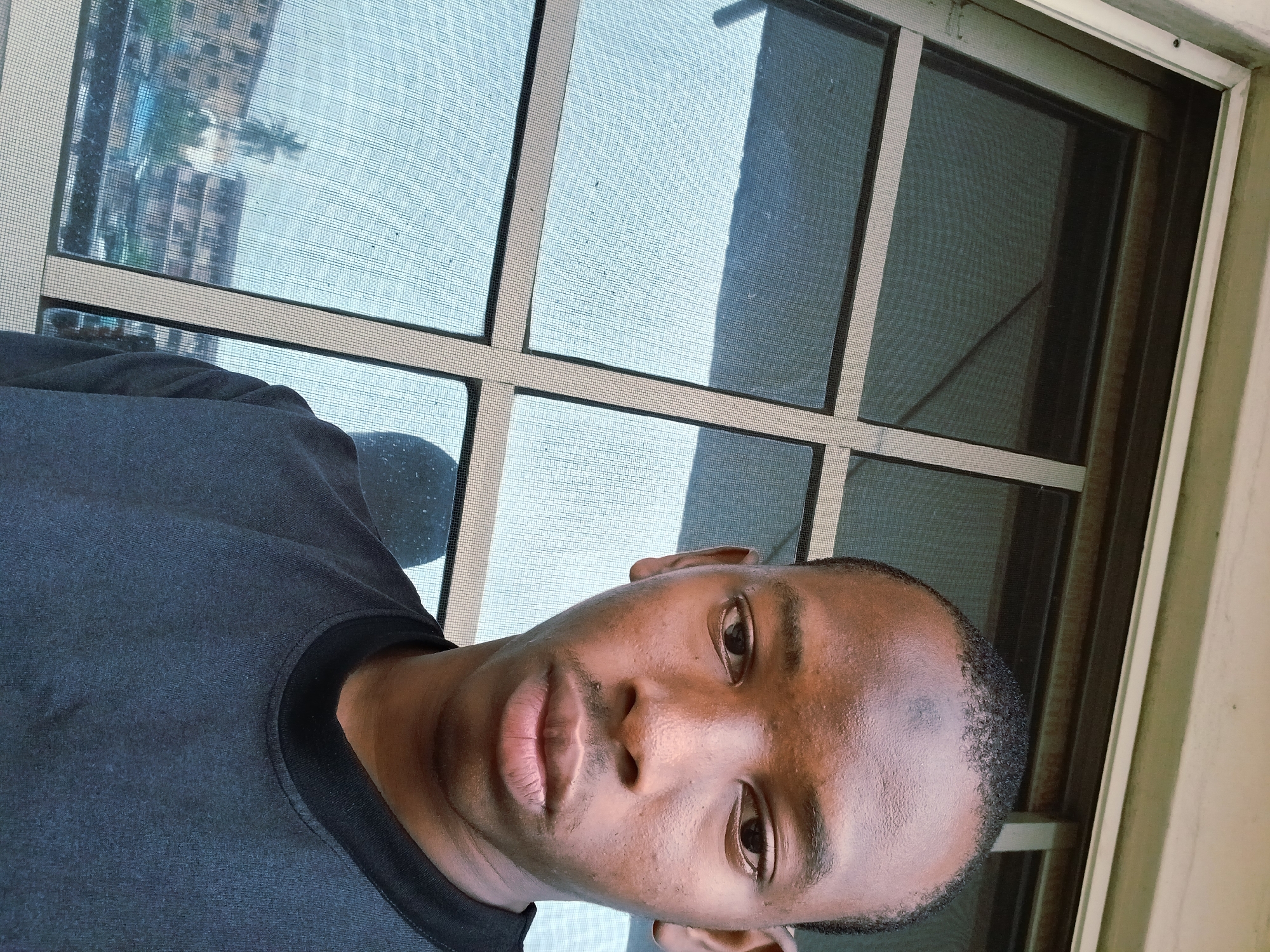 A young Black male is captured in a close-up selfie taken from a slightly low angle, positioned in front of a window. The background features a window with a grid of panes, some of which are covered by a fine mesh screen. Through the screen, buildings are visible, suggesting an urban environment. The lighting appears to be natural, coming from outside the window. The man is wearing a dark t-shirt. His expression is neutral, and he is looking directly at the camera. The overall scene suggests an indoor setting with a view of the outside.