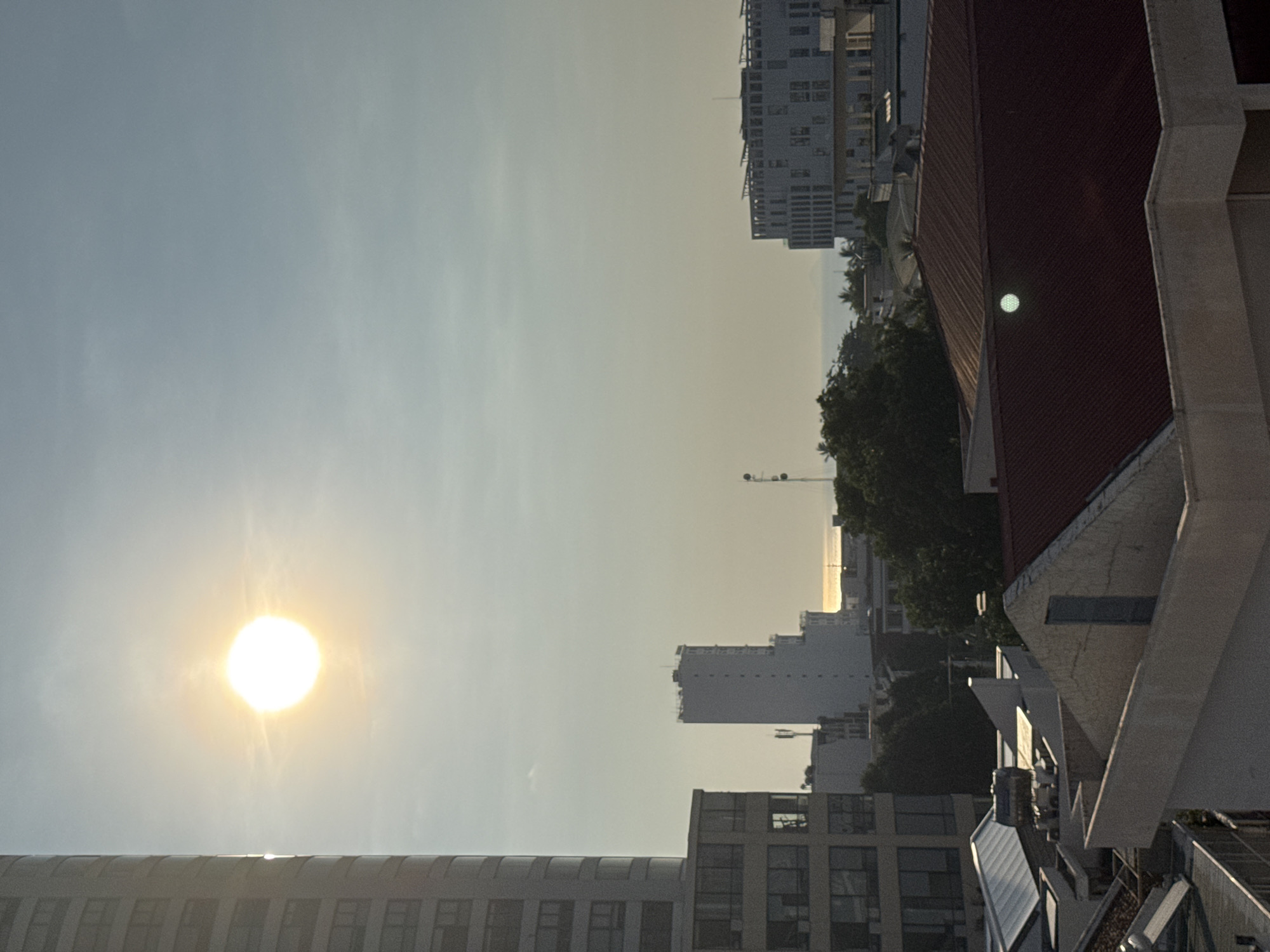 The image shows a cityscape with a bright sun in the sky. In the foreground, there are several rooftops of buildings, some with red tiled roofs. In the mid-ground, there are trees and other buildings, including a tall, slender white building and a modern-looking white building with many windows. In the background, there is a body of water, likely the ocean, with a faint horizon. The sun is prominent in the upper left quadrant of the image, casting a bright glow. This is a scene from the city of Nha Trang, Vietnam. There are no people visible in the image.