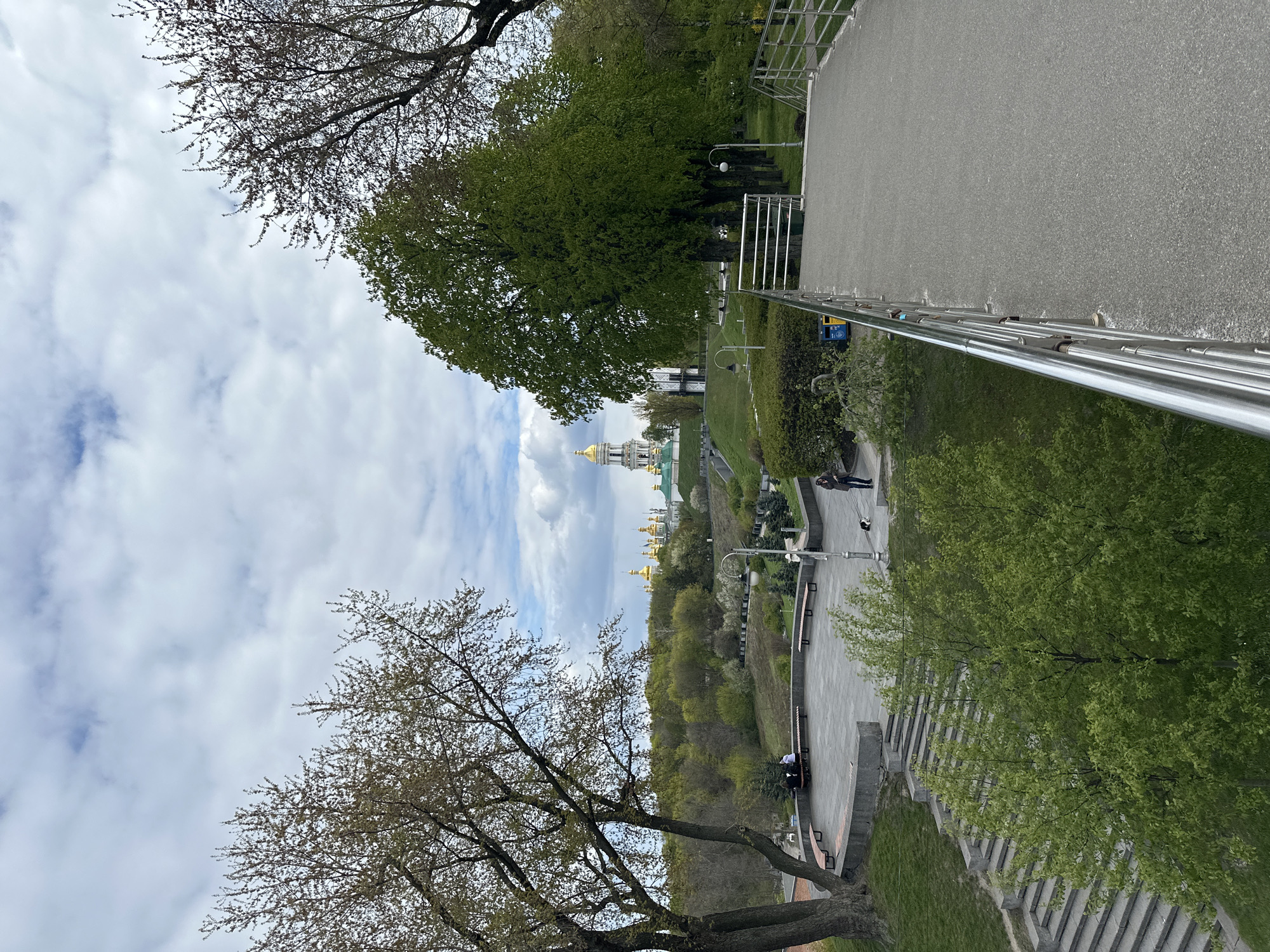 The image presents an elevated view of a park or promenade in Kyiv, Ukraine, under a cloudy daytime sky. A wide, paved pathway with a modern metal railing occupies the right side of the frame, extending towards the background. Several individuals are visible: one person stands near the railing on the upper path, another walks further along, and two people are seated on a bench on a lower, terraced area to the left.
The landscape is green, featuring numerous trees with varying degrees of foliage; some are fully leafed in bright green, while others display nascent leaves or sparser branches, suggesting late spring. Below the main promenade, the terrain slopes downwards, covered in grass and more trees, interspersed with concrete steps and benches.
In the mid-ground, prominent golden domes of a church or cathedral complex rise above the tree line, a recognizable feature of Kyiv's architectural heritage, such as the Kyiv-Pechersk Lavra. Streetlights are placed along the paths. A blue sign, with text that is unreadable, is attached to the metal railing. The scene conveys a peaceful public space, likely used for strolling, resting, and enjoying the view.