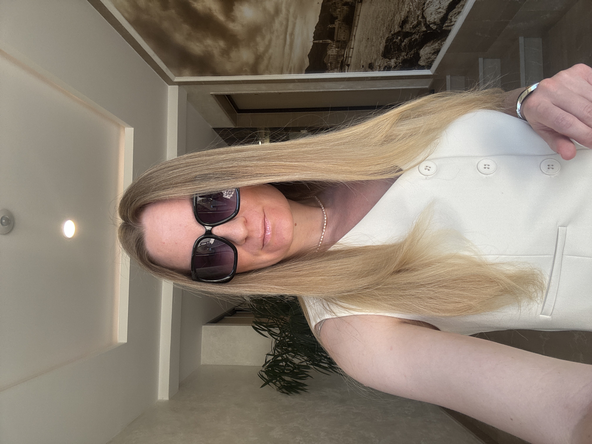 A close-up, sideways-oriented photograph captures a person, likely female, with long, straight blonde hair. She wears dark, square-framed sunglasses that obscure her eyes, and her lips are slightly parted in a faint smile, adorned with lip gloss. A delicate gold-colored necklace and a silver-toned cuff bracelet on her left wrist are visible. She is dressed in an off-white or cream-colored sleeveless top or dress, featuring three prominent round buttons down the front.
The setting appears to be a modern, well-lit interior, possibly within a hotel or upscale building in Budva, Montenegro. The background features light-colored walls with clean architectural lines and recessed lighting. A striking, sepia-toned panoramic mural or ceiling panel depicting ancient stone structures or ruins against a dramatic sky is visible overhead. Below, parts of a dark green, leafy indoor plant are visible, along with hints of light-colored flooring and structural elements that suggest a multi-level interior or staircase. The lighting is bright and even, consistent with daytime indoors. No visible text is present in the image.