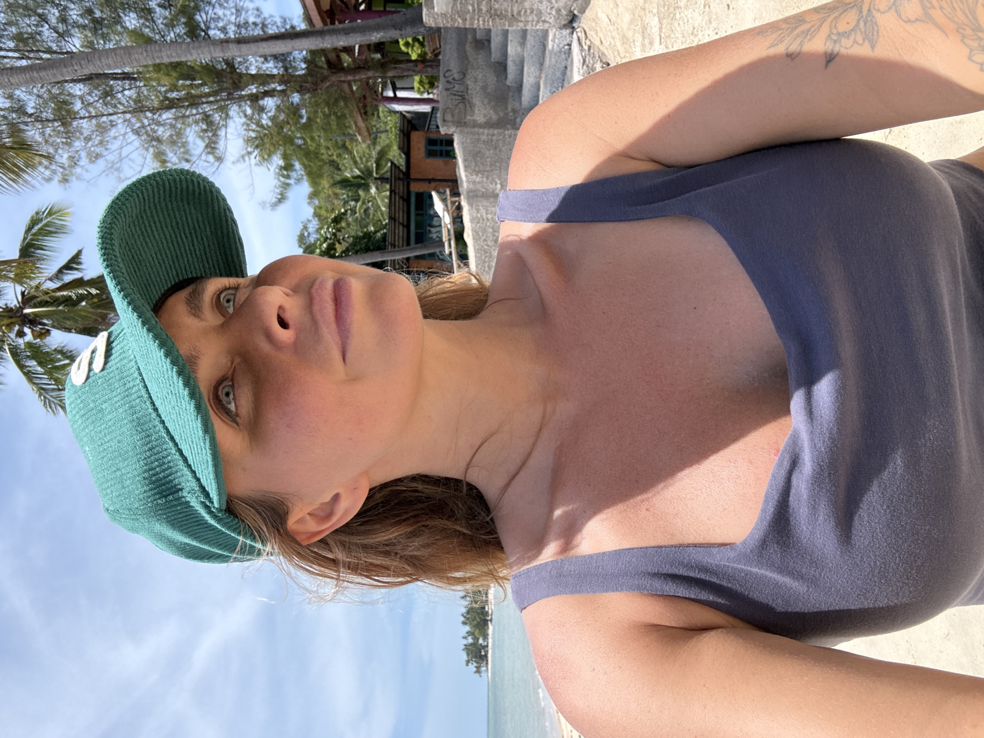 A woman wearing a teal baseball cap and a dark blue tank top is photographed outdoors on a sunny day. She is looking up and to the right, with her mouth slightly parted. Her hair is light brown and falls around her shoulders. In the background, there is a beach with sand, palm trees, and the ocean. Further back, there are buildings with colorful roofs and a concrete staircase. The image appears to be taken in a tropical location.