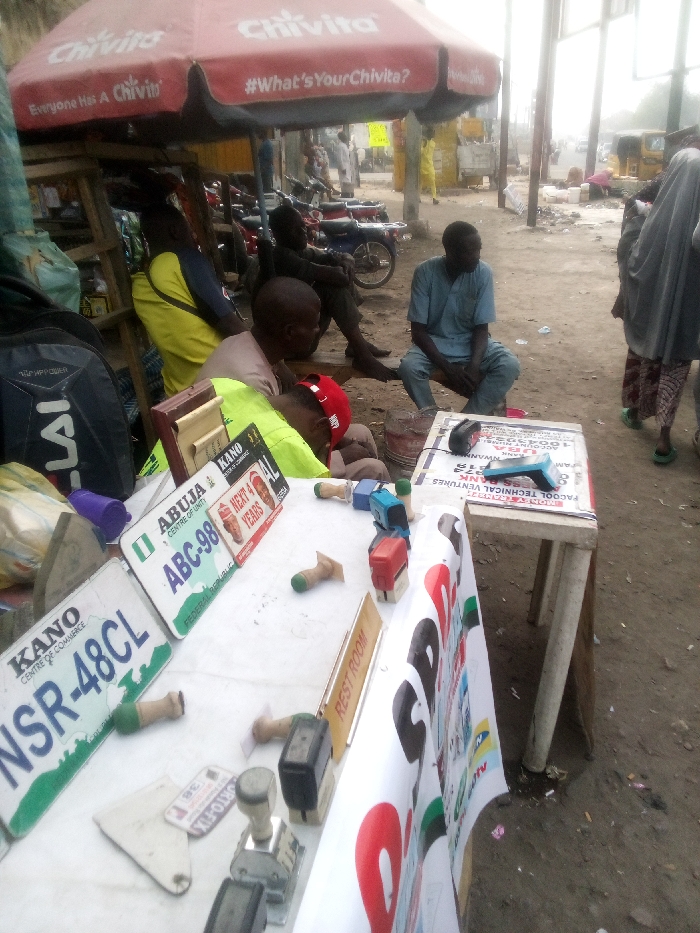 The scene depicts an outdoor street vendor stall in Sabon Gari East, Nigeria. Underneath a red Chivita-branded umbrella, various individuals are present. A man in a light blue tunic is seated on a low stool to the right of a white table display, looking towards the right. Another individual in a light brown shirt and red cap is positioned over the table, obscured, suggesting leaning or sleeping. A third person, wearing a yellow and blue shirt, is seated or leaning near a black backpack to the left. Behind them, another man sits on a bench in darker attire, and multiple motorcycles are parked.
The white table, supported by wooden trestles, serves as the primary display. On its surface are two Nigerian vehicle license plates: one green and white for "ABUJA ABC-98 FEDERAL REPUBLIC" and another white and green for "KANO NSR-48CL". Numerous hand stamps, including self-inking and traditional wooden-handled types, are scattered across the table. A small "REST ROOM" sign and a black electronic device are also present. The front of the table is draped with a white banner displaying text and logos, including "FACOOL TECHNICAL VENTURES" and a "UBA" bank logo. The ground is unpaved, appearing as dirt or loose earth. In the background, an unpaved road extends, populated by additional individuals, structures, and a tricycle taxi, under hazy daylight conditions. A person in light-colored religious attire walks away from the camera on the far right.