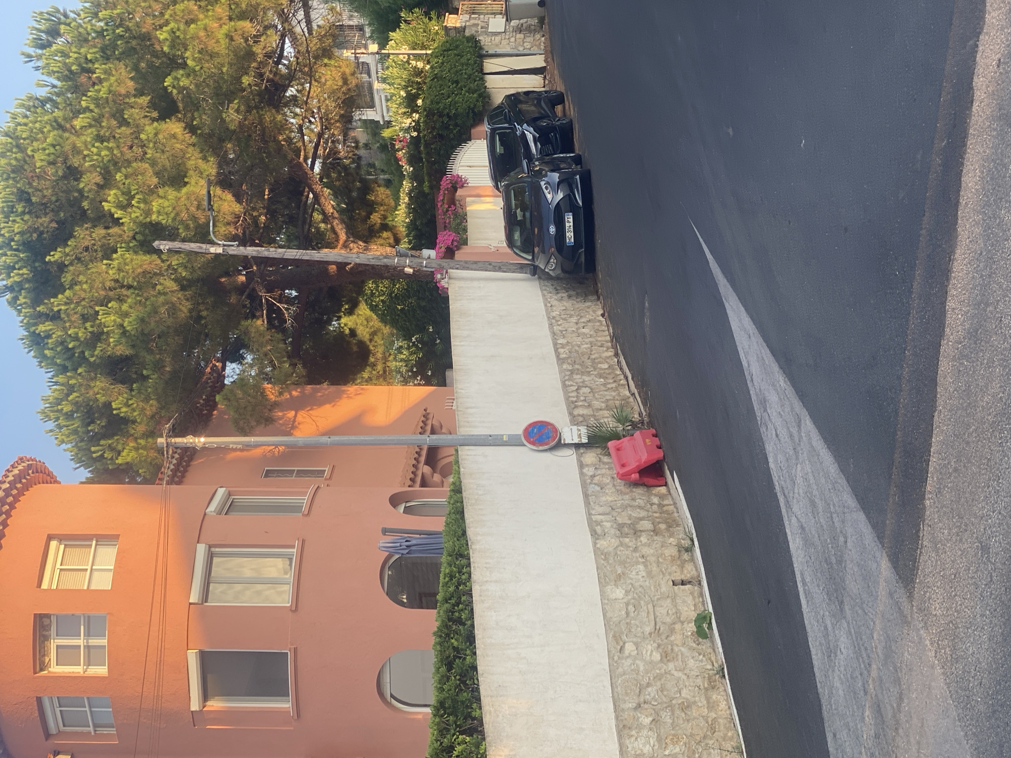 The image shows a street scene in Antibes, France, on a sunny day. The foreground features a dark asphalt road with a white directional arrow painted on it, indicating a turn. To the left of the road, a light-colored stone wall borders a sloped area, topped by a white rendered wall. Behind the white wall, a pink building with arched windows and a tiled roof is visible. A tall, slender lamppost stands beside the white wall.
Further back, a large, mature pine tree dominates the upper right portion of the image, its branches casting shadows. In front of the white wall, two dark-colored cars are parked along the side of the road: a smaller hatchback on the left, displaying a French license plate "MC 314 PY", and a larger SUV to its right. Two red plastic barriers are placed near the base of the white wall. Pink and white flowering bushes are visible behind the cars and the white wall. The sky is clear and blue.