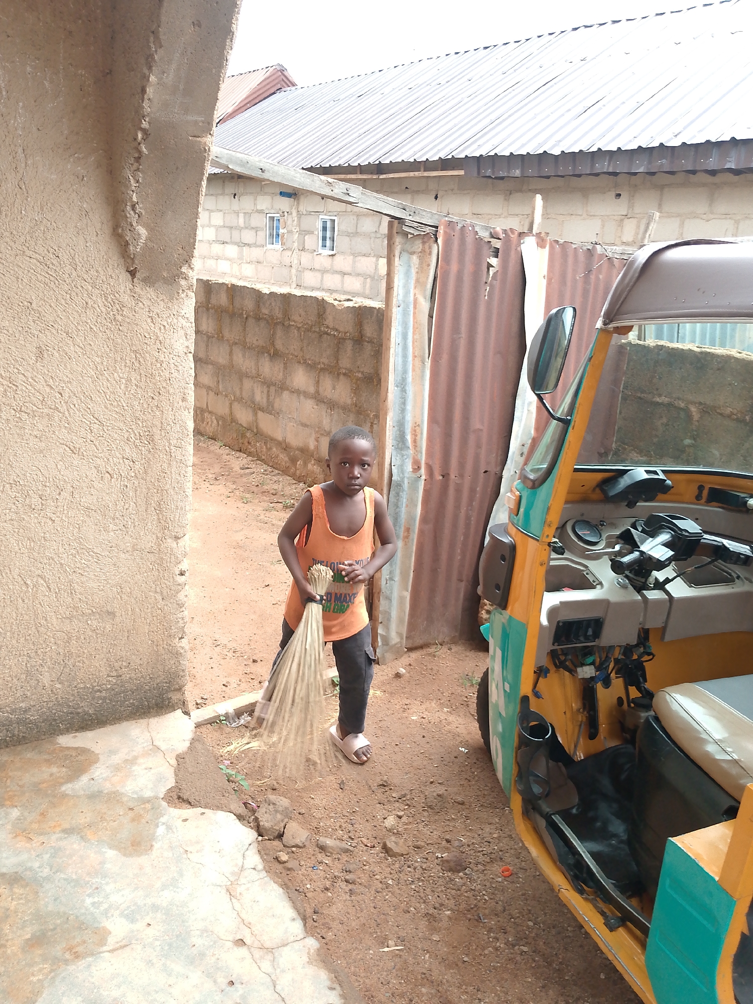 A young child in an orange tank top and dark pants sweeps the ground near a yellow and green auto-rickshaw, commonly known as a "keke," in Jos, Nigeria. The child holds a bundle of broom grass. The background includes a concrete wall, a brick building with windows, and corrugated metal fencing. The ground is dirt and partially paved.