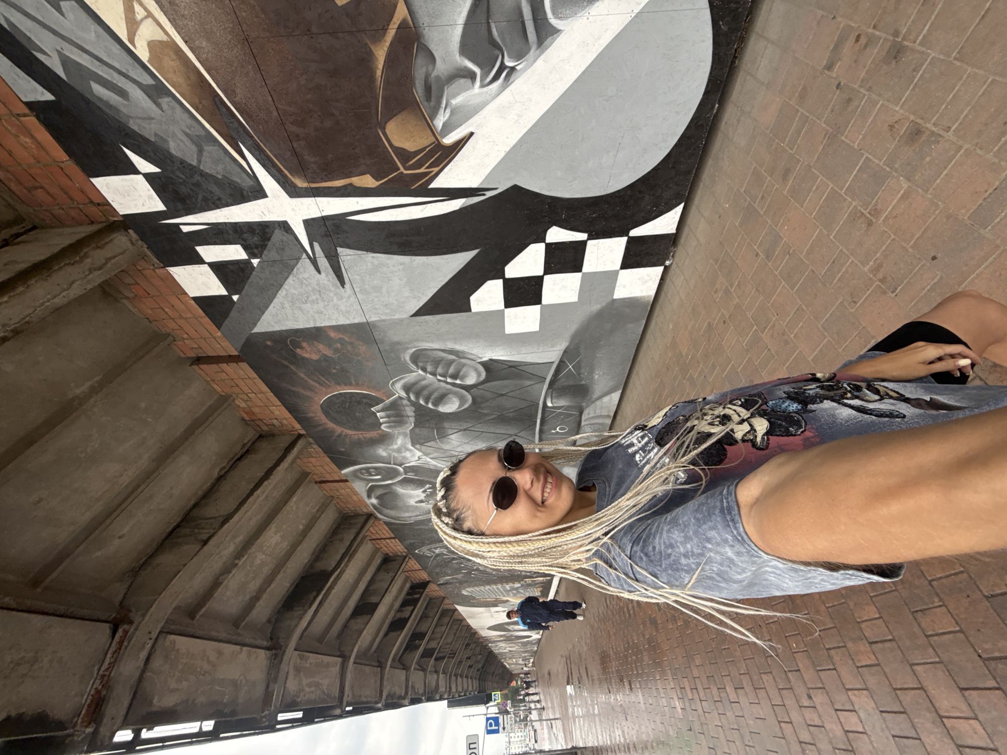 A young woman with blonde braids and sunglasses is taking a selfie in an urban setting. She is smiling and wearing a blue t-shirt with a graphic print and denim shorts. The background features a large mural with abstract designs, checkerboard patterns, and what appears to be a stylized eye. To her left, there is a concrete structure with arched supports, and to her right, a brick pathway. In the distance, a street with buildings and traffic signs is visible. A man in a blue jacket is walking away from the camera in the background. The scene appears to be outdoors, possibly during the daytime, with overcast weather. The location is identified as Rusanovo, Russia. Visible text includes road signs with Cyrillic letters and a "P" symbol, indicating parking.