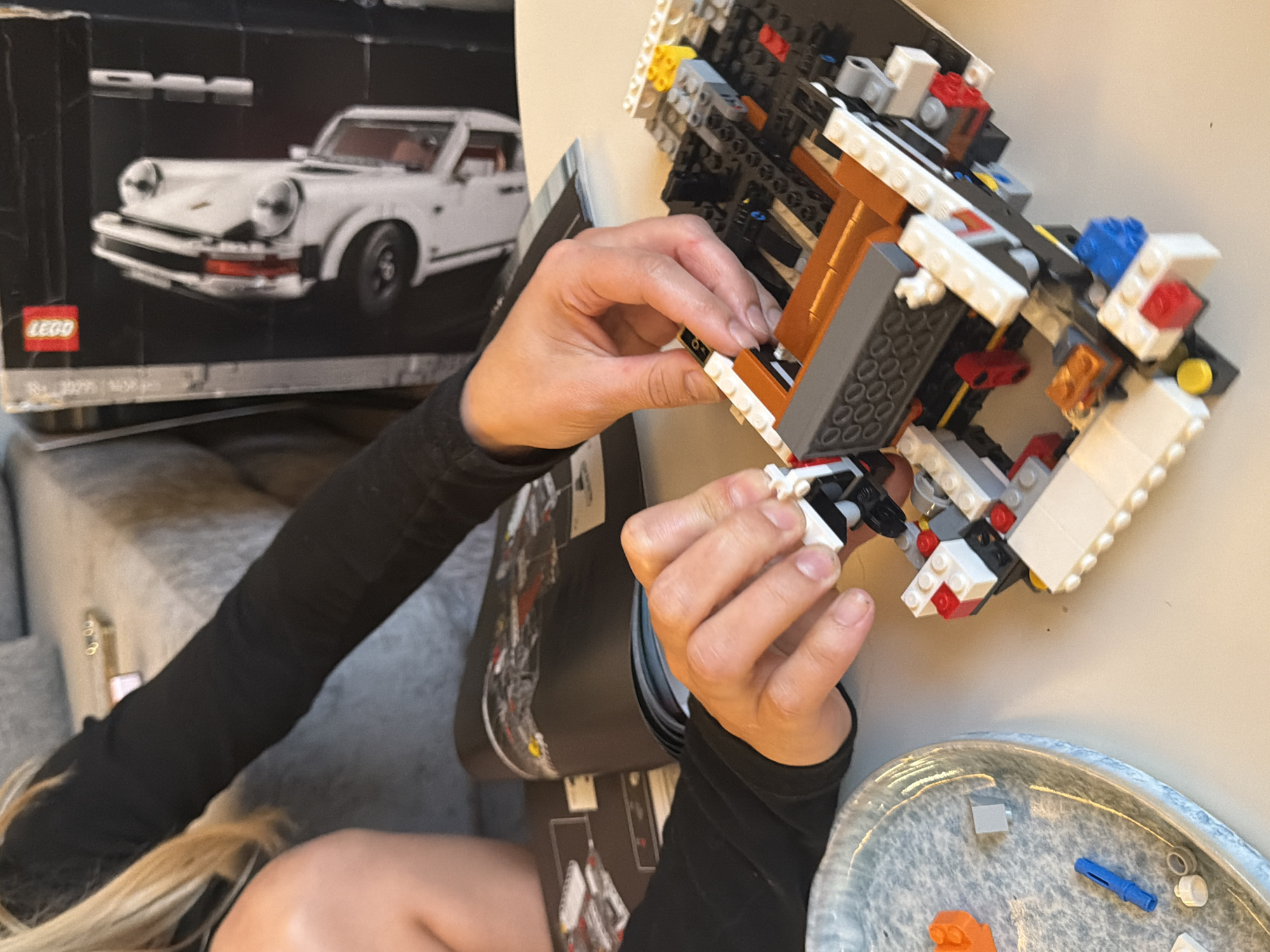 A woman with blonde hair, wearing a long-sleeved black shirt, is building a complex Lego structure with white, gray, black, orange, and red pieces. The project appears to be related to a Lego Porsche 911, as the box for that model is visible in the background. Lego instruction manuals are also present. A dish containing loose Lego pieces sits nearby. The scene takes place indoors. There is no information available in the image to indicate the location in the city of London, United Kingdom.