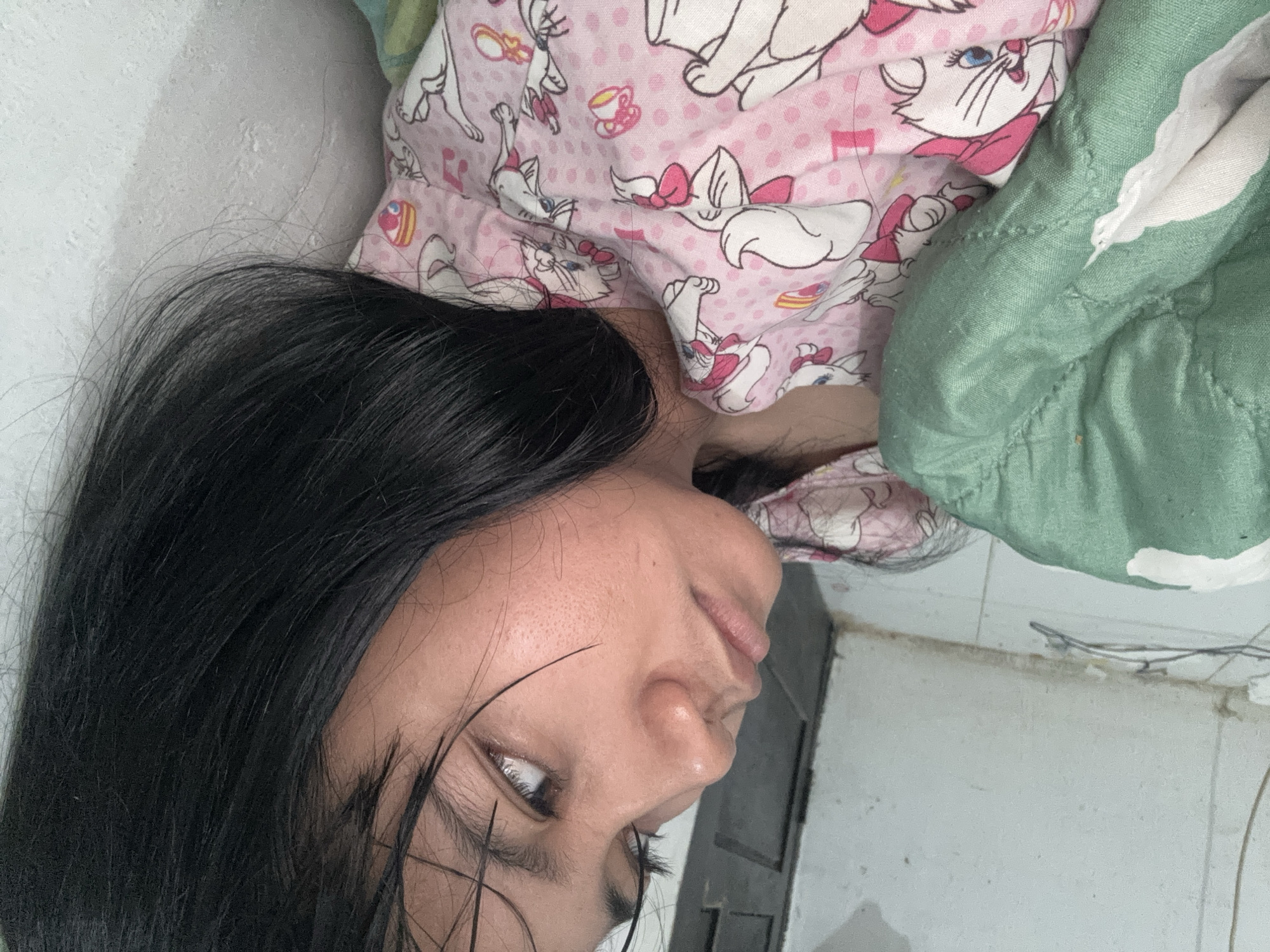 A young person lies in bed, covered by a green blanket and wearing pajamas featuring cartoon cats. The scene is indoors, with a white tiled floor and a textured white wall visible. The person's head is turned to the side, looking upwards. There are wires visible on the floor near the wall. The lighting suggests an indoor setting, possibly during the day or with artificial light. The overall impression is one of a domestic environment.